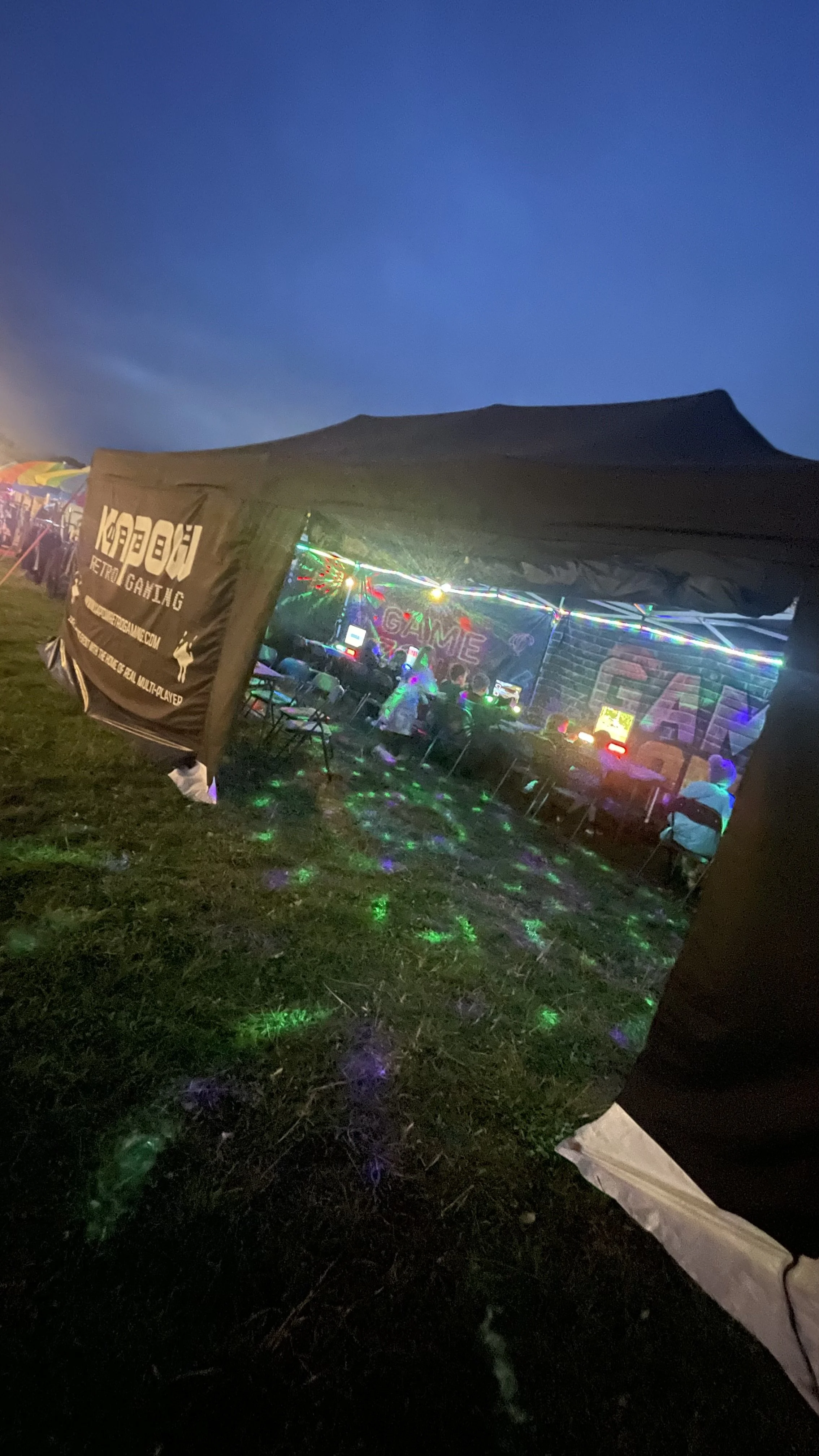 An outdoor gaming event under a tent with LED lights and people sitting at tables, with a dark evening sky in the background.
