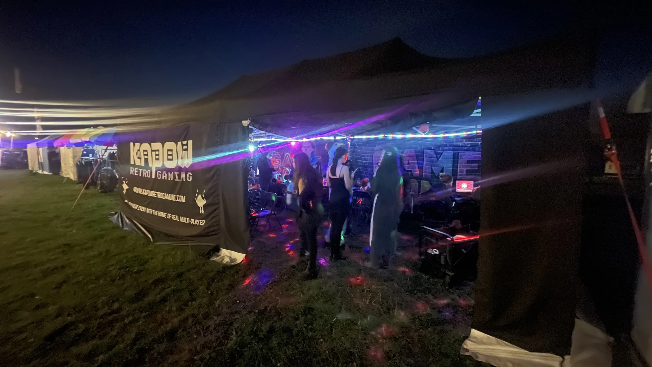 Nighttime scene inside a gaming tent with neon lights, people gathered around gaming computers, and a banner advertising 'KAPOW! RETRO GAMING' outside the tent.