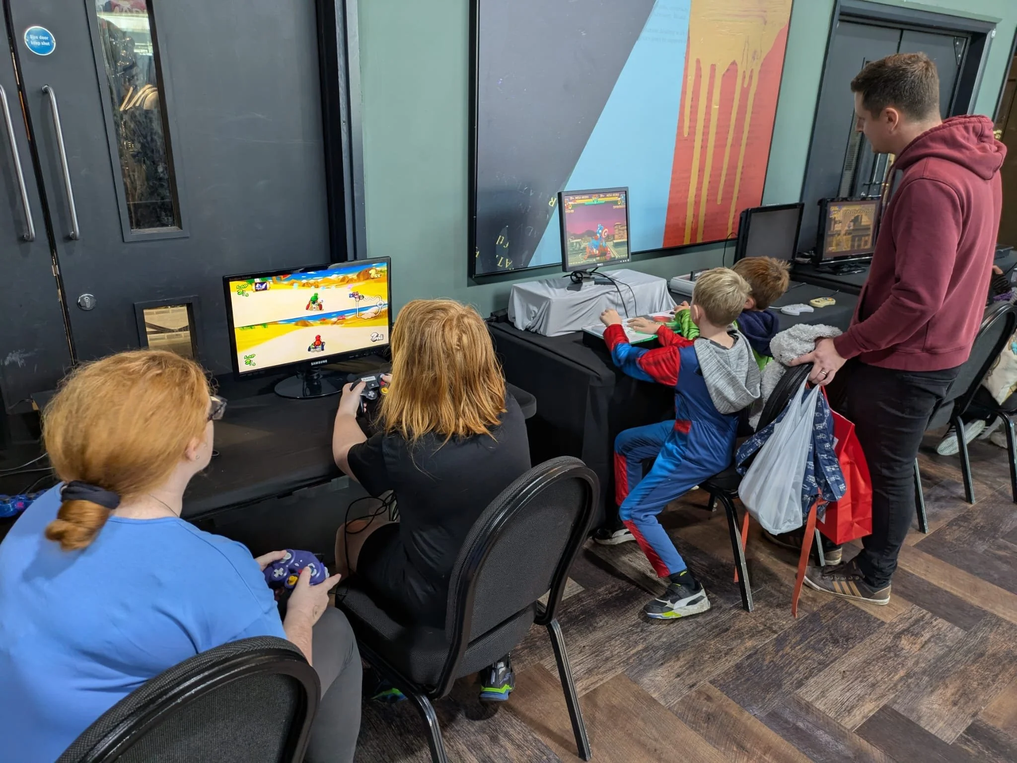 Children and an adult playing video games on multiple gaming consoles set up along a row of tables in a gaming center or arcade. The children are sitting in chairs, focused on screens showing racing games, with the adult supervising or watching.