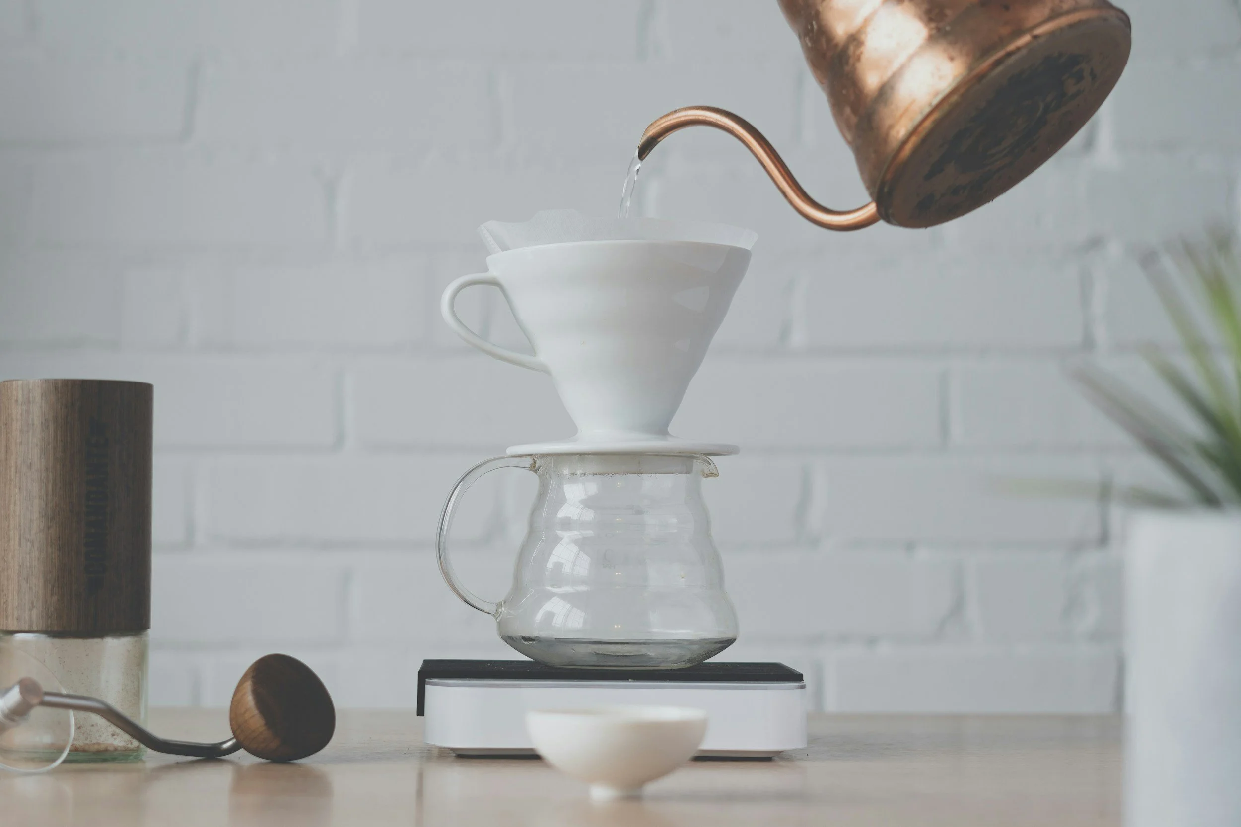 Pour-over coffee setup with water being poured from a gooseneck kettle into a ceramic dripper on a glass carafe, placed on a digital scale, with a grinder and a small bowl nearby.