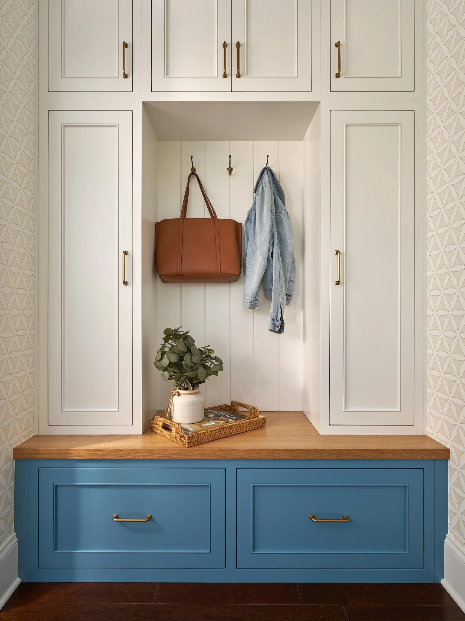 Blue and white custom mudroom cabinetry in Maryland with concealed storage, brass pulls, coat hooks, and a natural wood bench.