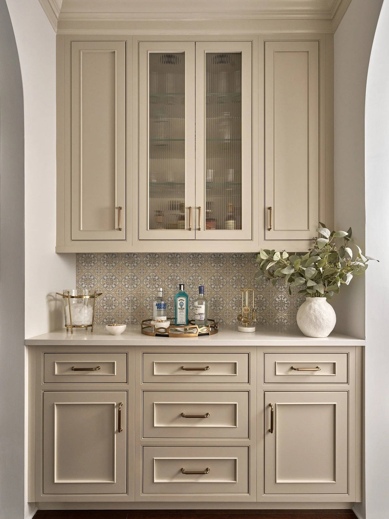 Built-in home bar in MD with taupe cabinetry, reeded glass doors, patterned tile backsplash, brass hardware, and a white countertop.