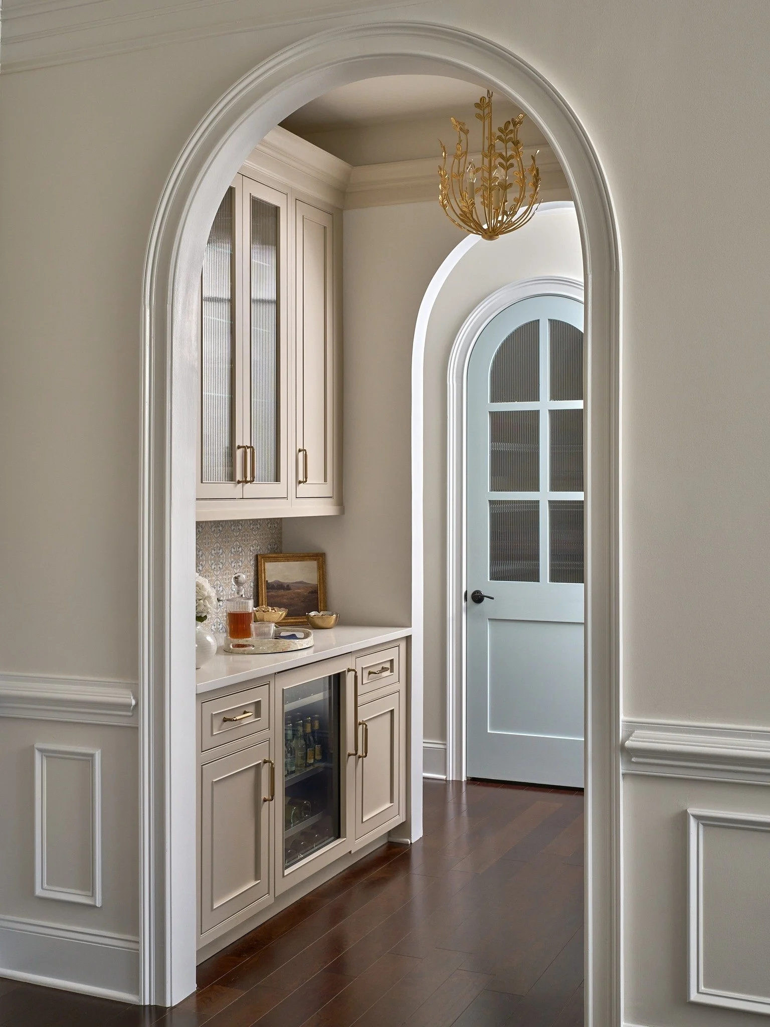 Custom Maryland butler’s pantry framed by an arched opening with taupe cabinetry, brass hardware, reeded glass, and a pale blue arched door.