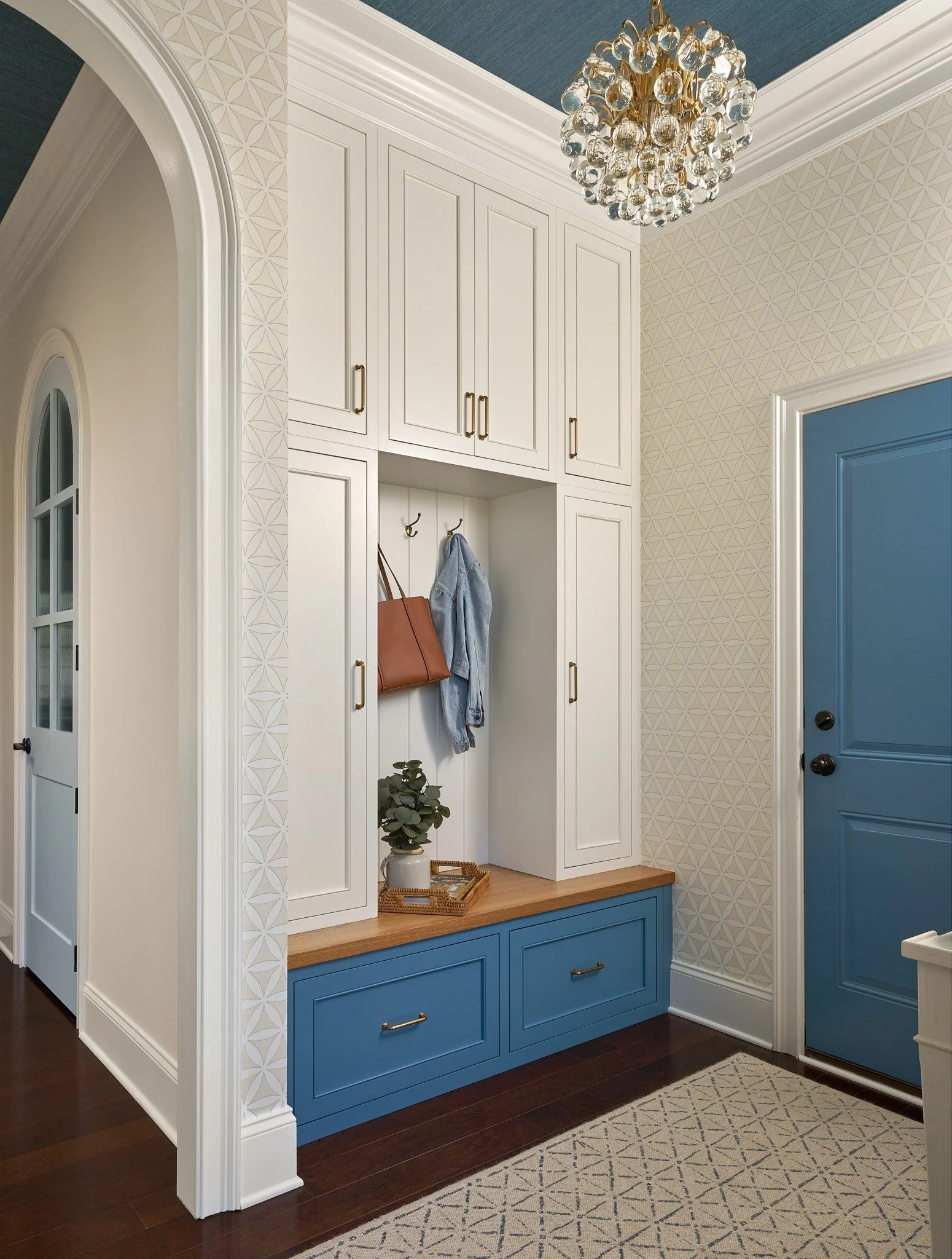 Blue and white mudroom in MD featuring custom cabinetry, a built-in bench, patterned wallpaper, a crystal chandelier, and arched trim.