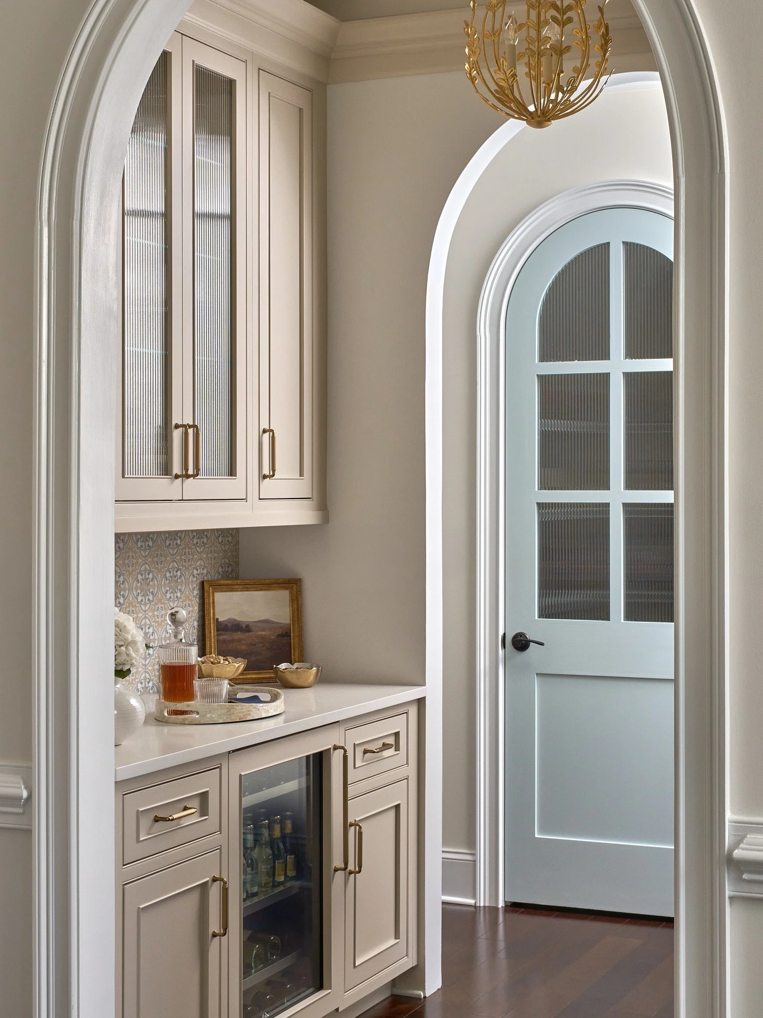 Arched butler’s pantry in MD with taupe cabinetry, reeded glass doors, a beverage refrigerator, patterned tile, and brass lighting.