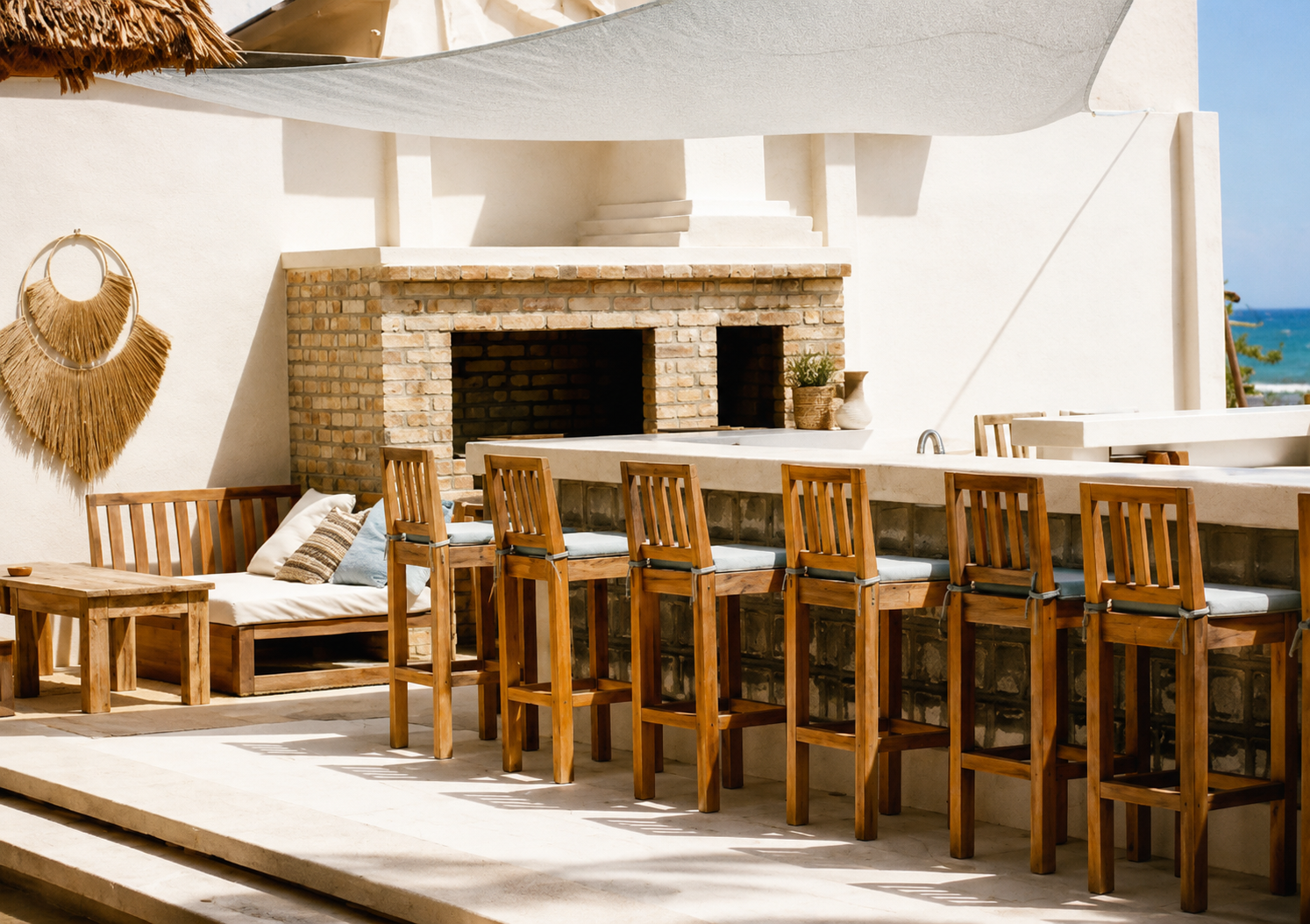 Mediterranean outdoor kitchen with bar seating, brick fireplace, wood furniture, shade sail canopy, and coastal-inspired design.