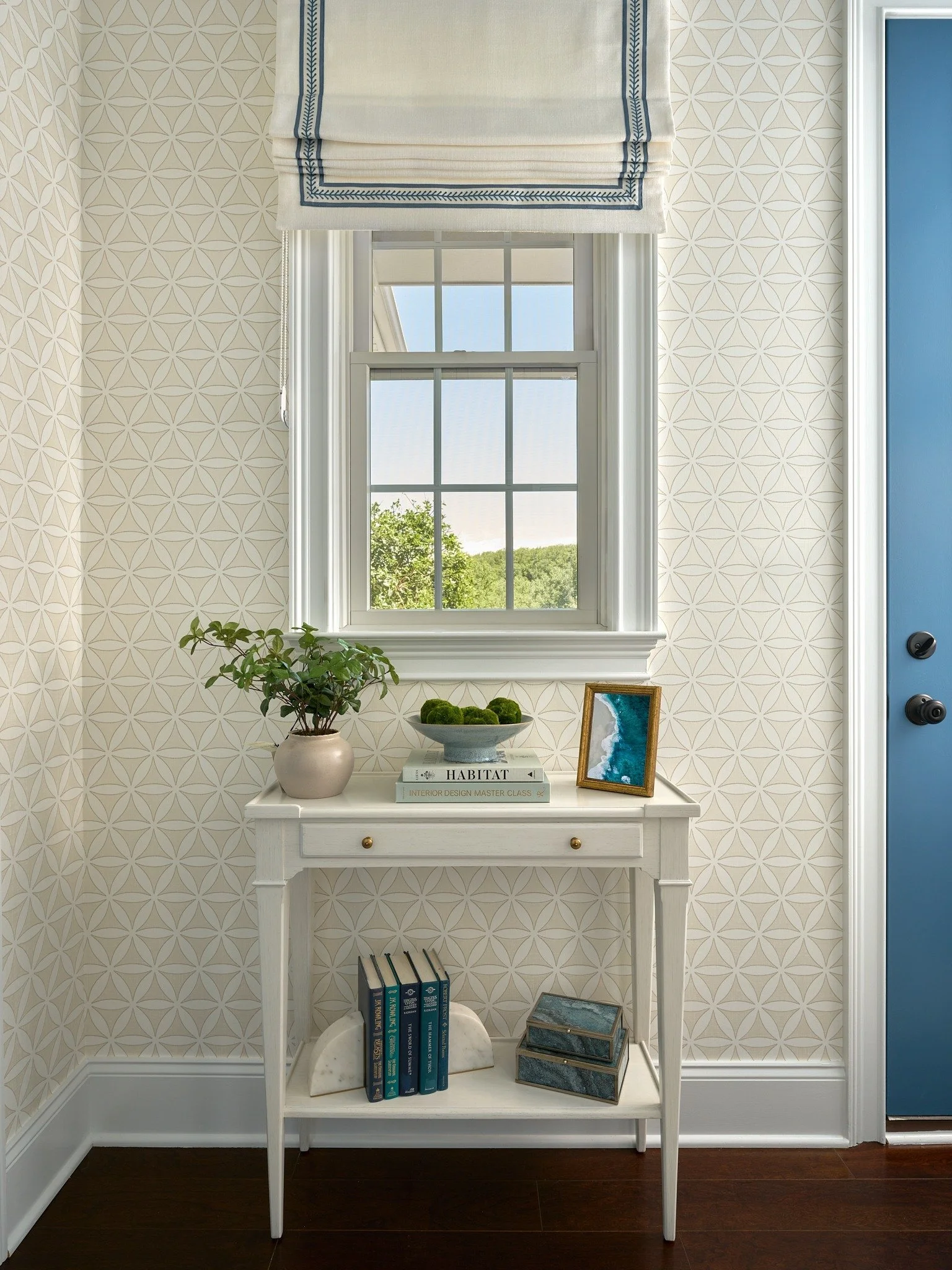 Maryland entryway with geometric wallpaper, a custom Roman shade, blue door, and white console table styled with books and greenery.