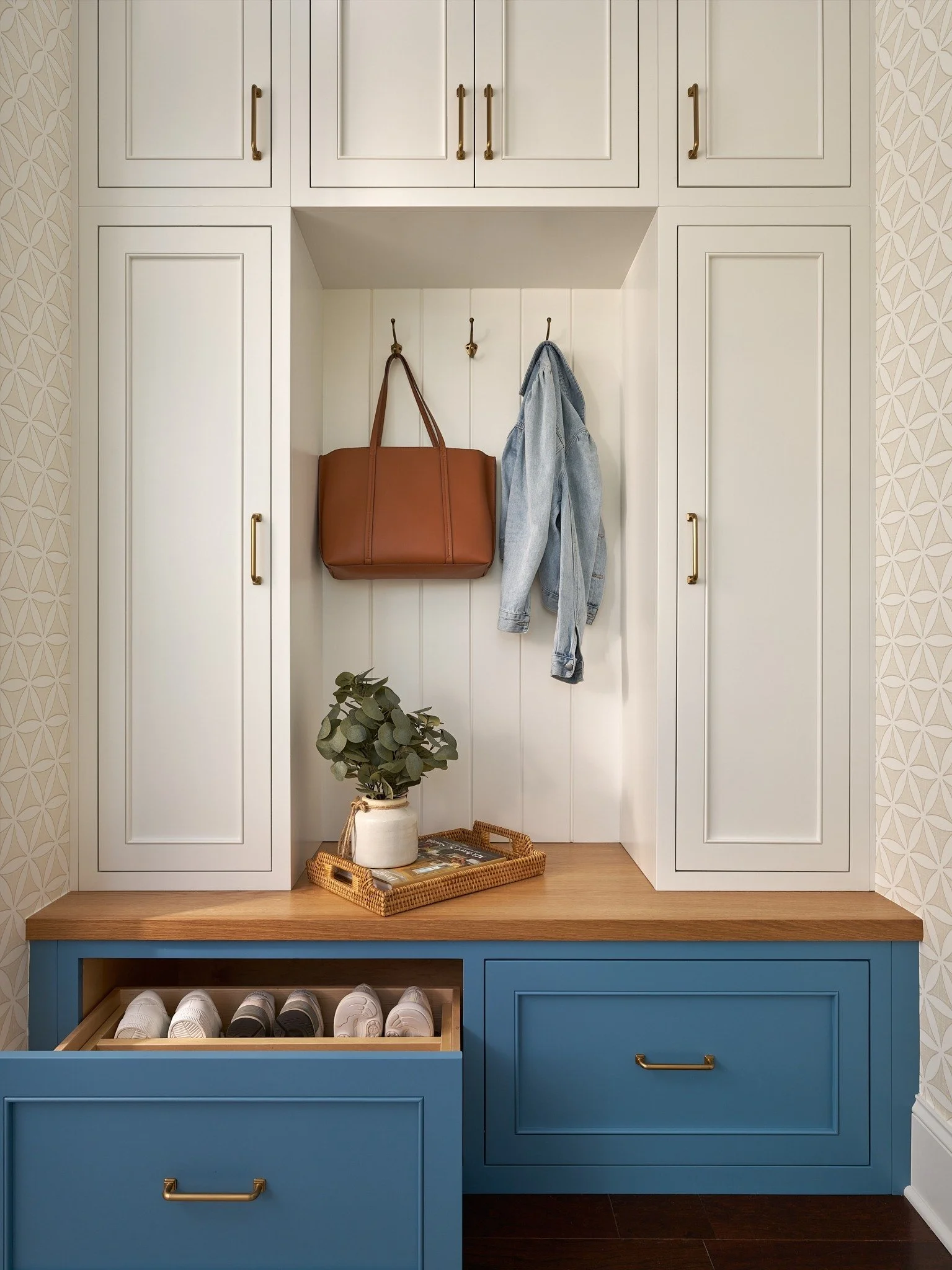 Custom mudroom built-ins in MD with white cabinets, blue shoe-storage drawers, brass hardware, coat hooks, and a wood bench.
