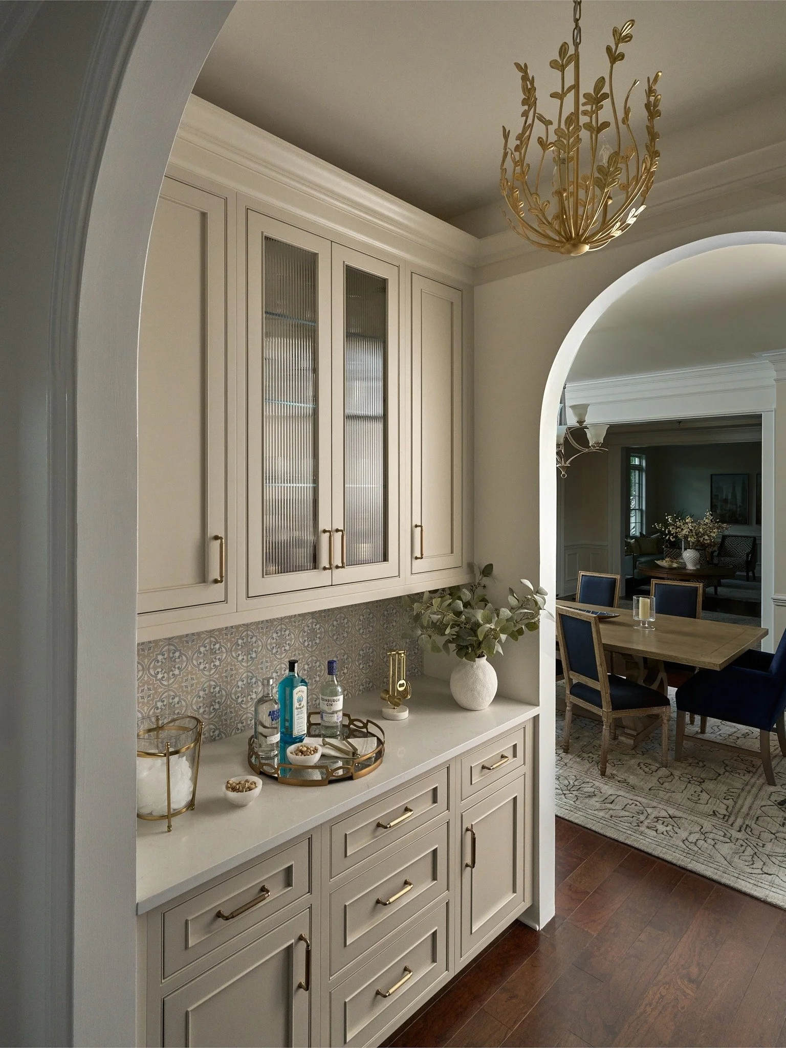 Maryland home bar with taupe cabinetry, reeded glass, patterned tile, and brass lighting beside an arched opening to the dining room.