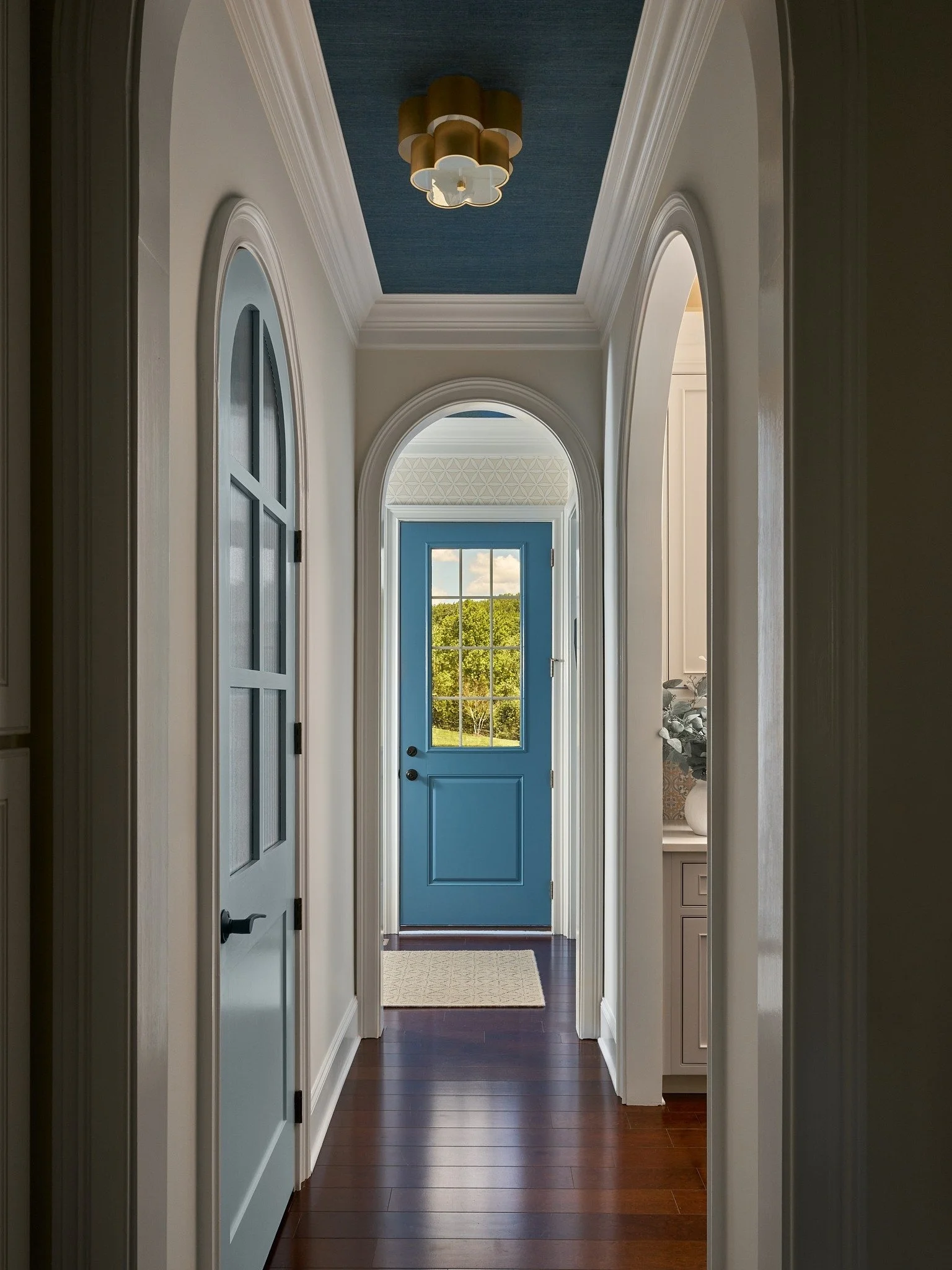 Maryland hallway with arched doorways, a blue paneled door, dark wood floors, a blue grass cloth ceiling, and sculptural brass lighting.
