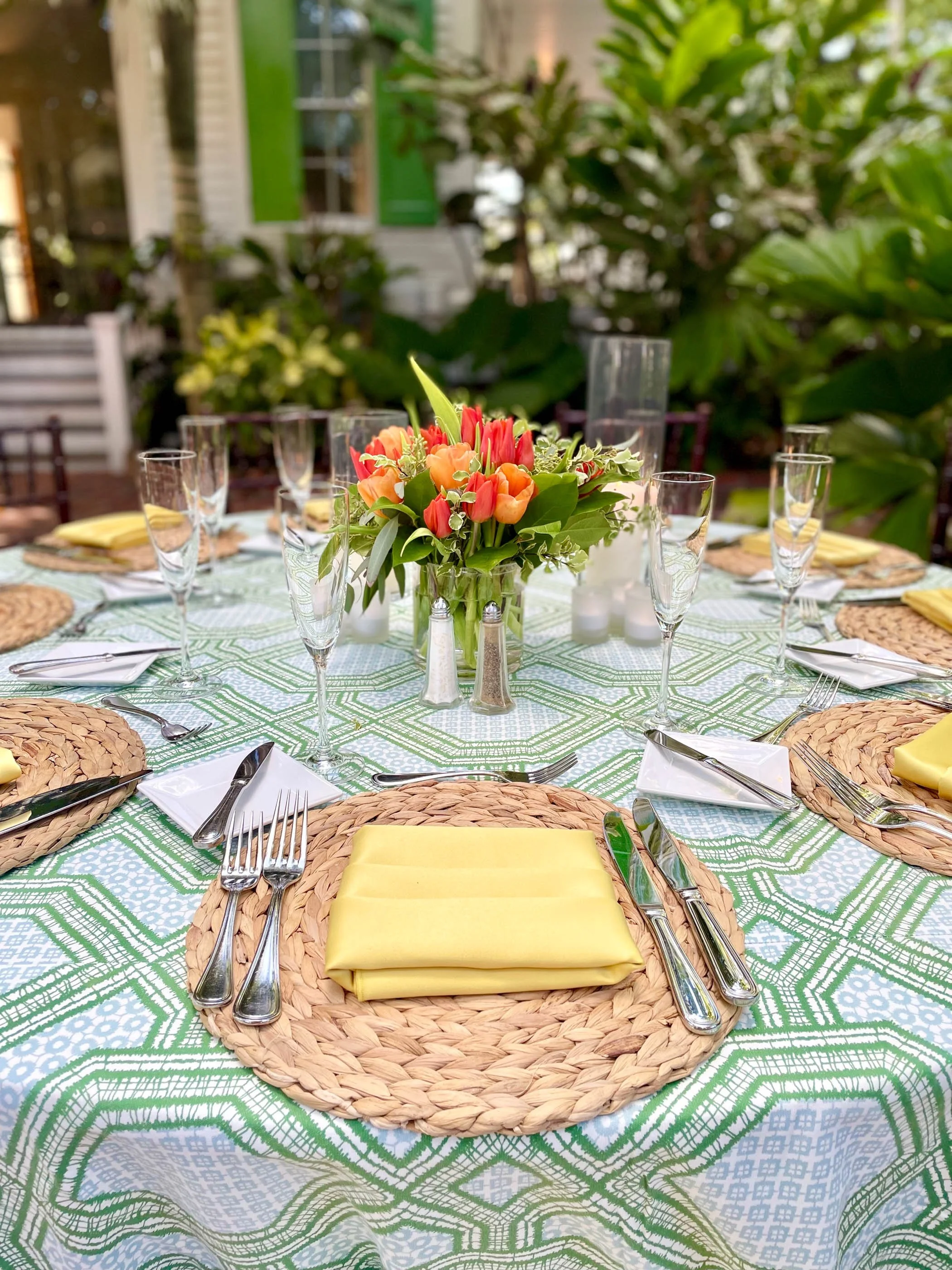 A round outdoor dining table set with woven placemats, yellow napkins, and silverware, centered with a floral arrangement of pink and orange flowers, on a green and white patterned tablecloth surrounded by greenery.