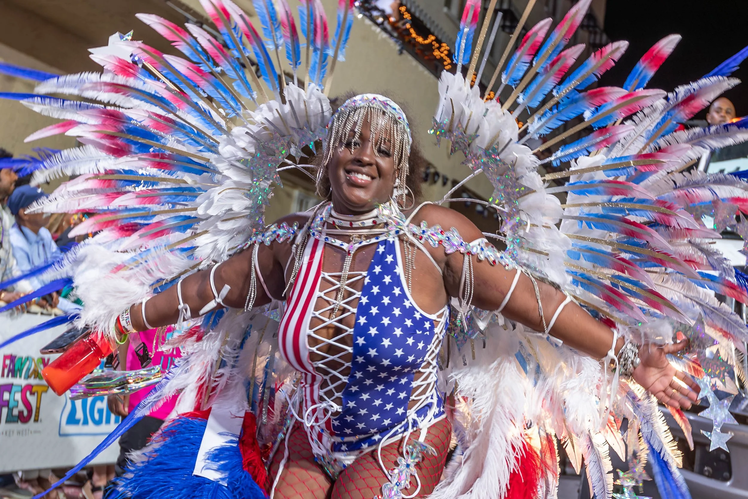 A woman dressed in a colorful, patriotic costume with red, white, and blue stars and stripes, adorned with large feathers and rhinestones, celebrating at a parade.