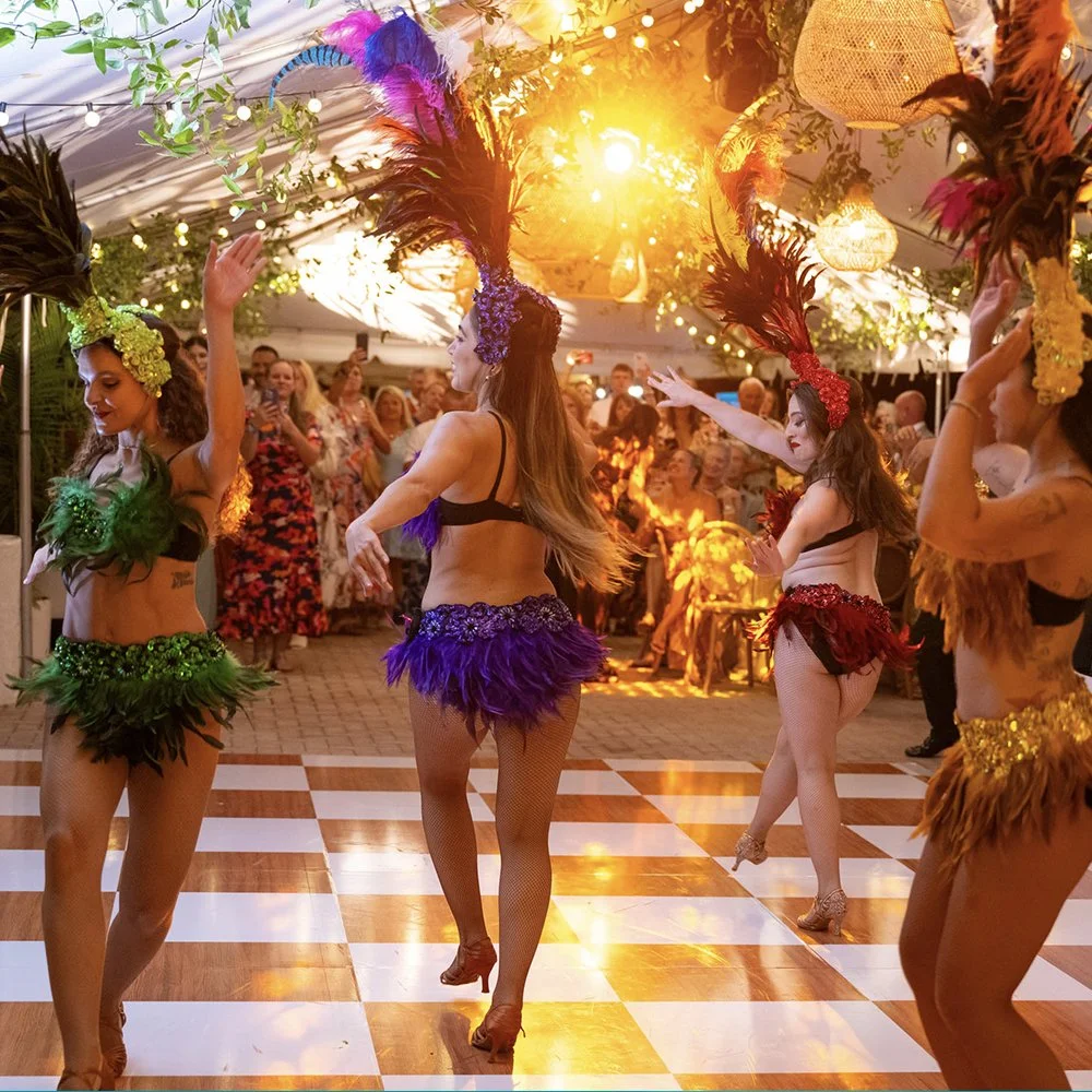 Hula dancers performing on a checkered dance floor under a decorated tent with audience watching