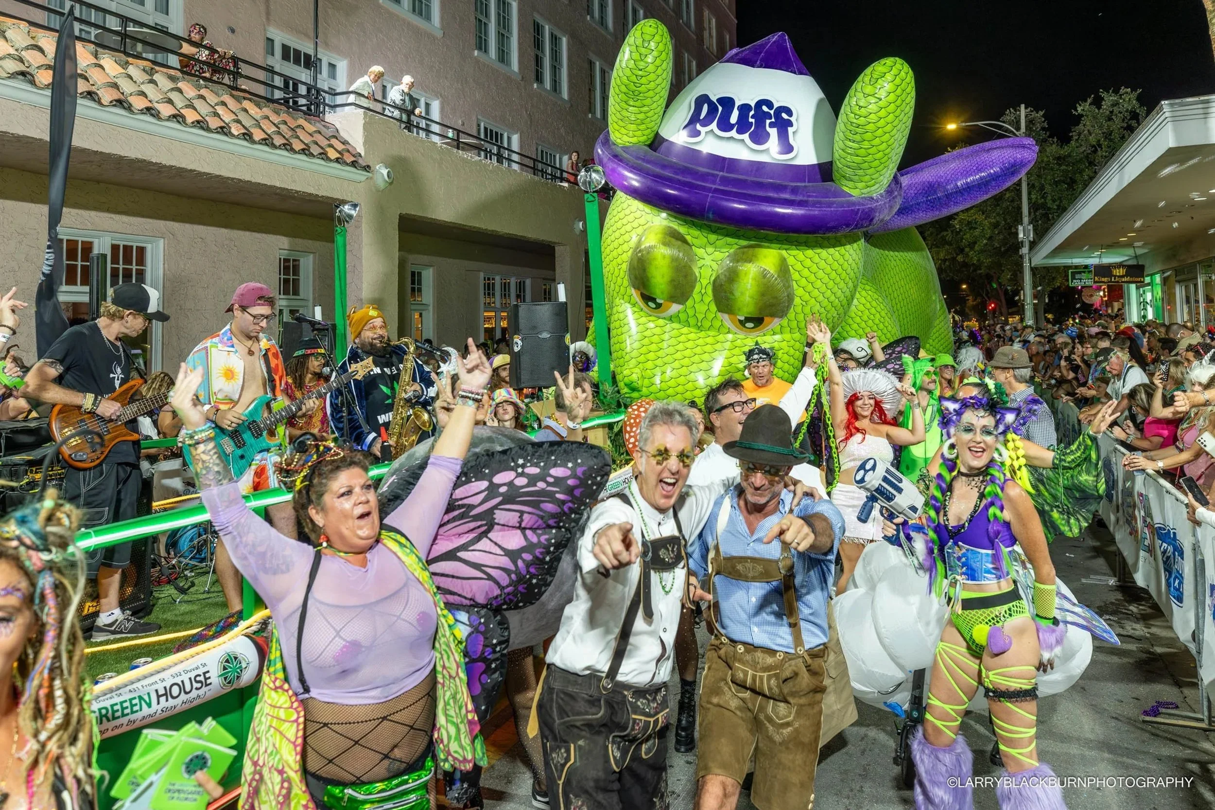 A lively street parade at night with costumed performers, a large inflatable dragon, a band playing, and spectators watching from balconies and sidewalks.