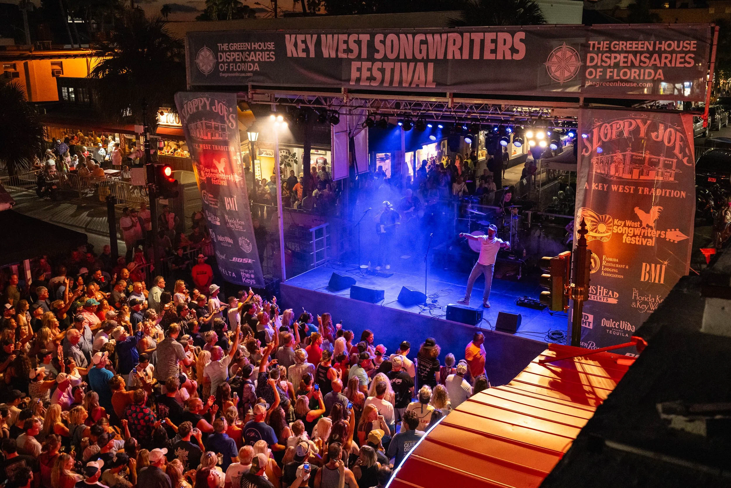 A crowd of people watching a live performance on an outdoor stage at night during the Key West Songwriters Festival. The stage has a banner with event sponsors and is lit with colorful lights, with a singer performing and audience members taking pict