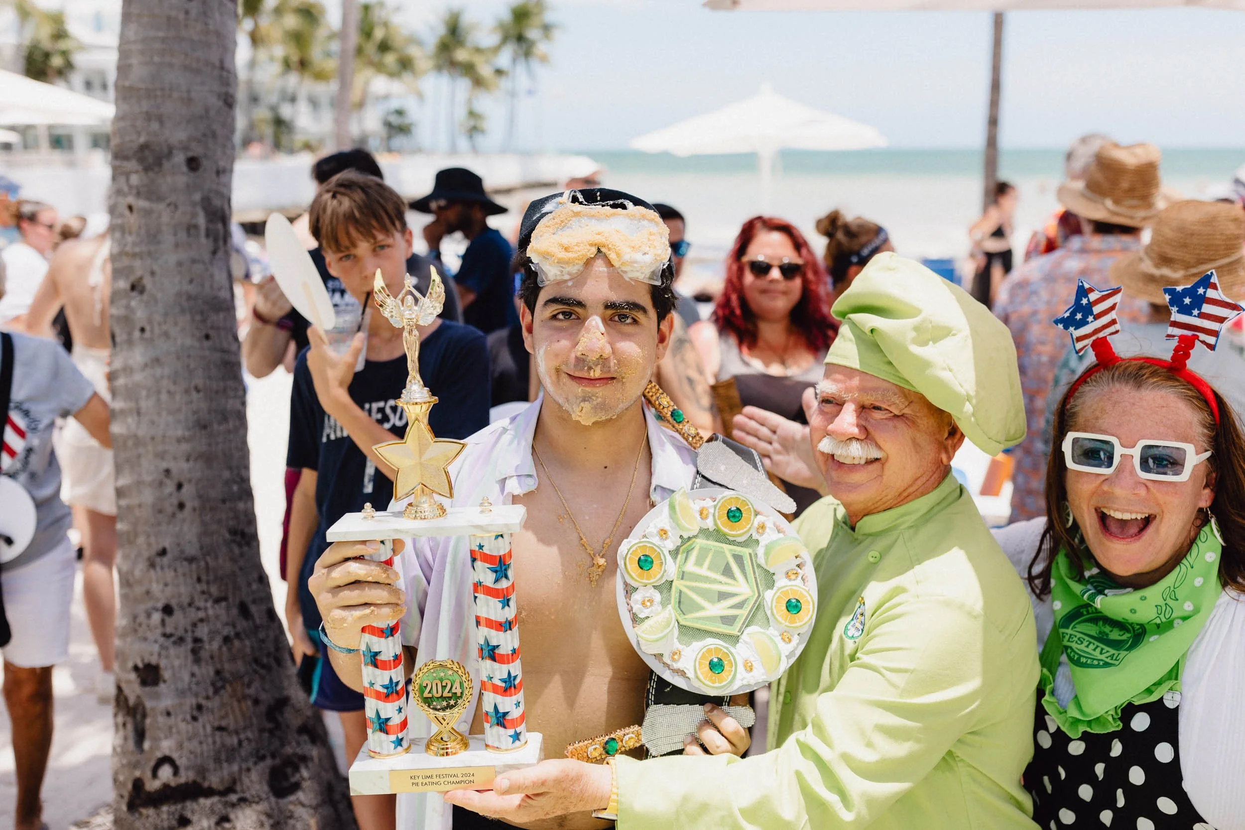Young man holding trophy with cake on his head, celebrating at a beach event, surrounded by smiling people with patriotic accessories, with the ocean and palm trees in the background.