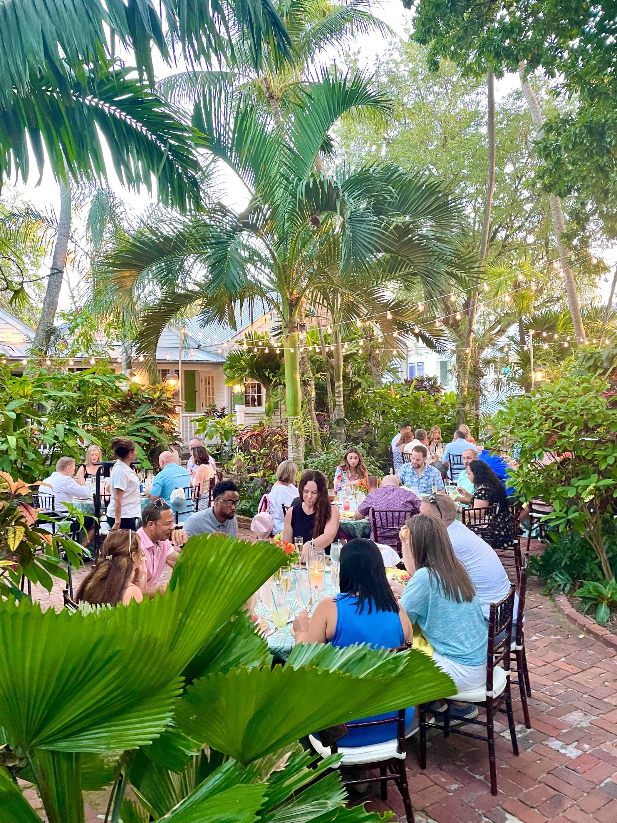 People dining at an outdoor tropical garden restaurant during the evening, surrounded by lush green plants and string lights.