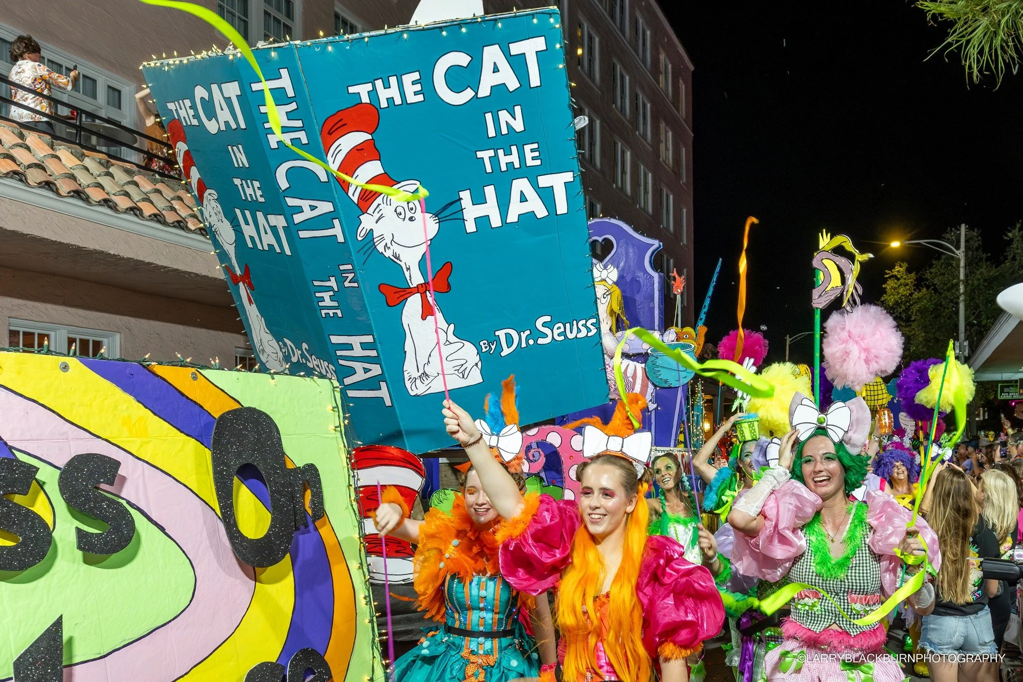 Colorful parade featuring people in vibrant costumes, some with wigs and face paint, celebrating in front of a float decorated with a large banner of 'The Cat in the Hat' by Dr. Seuss, illuminated at night.