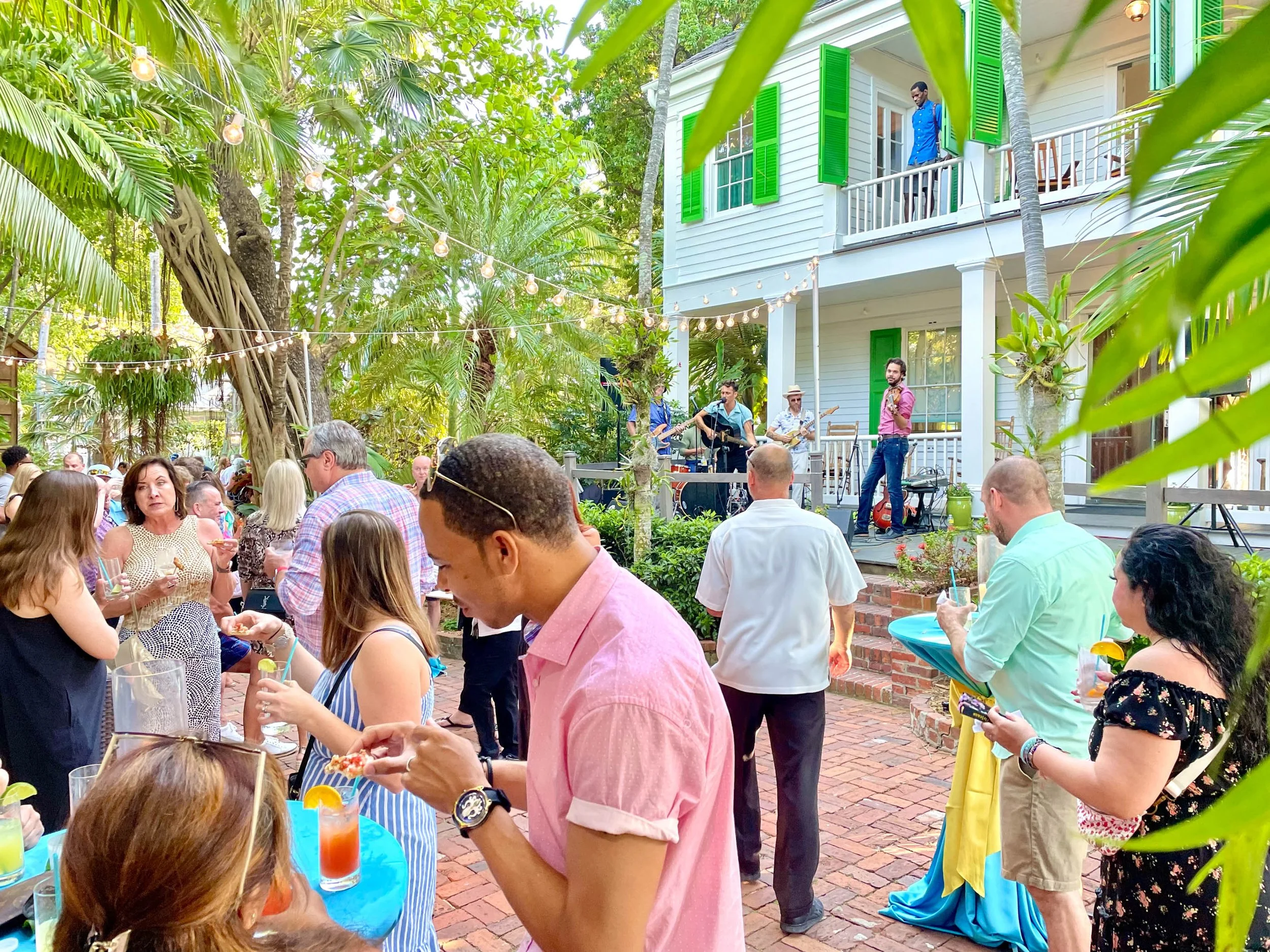 A lively outdoor party with people socializing, drinking, and enjoying live music on a patio surrounded by lush greenery and string lights, with a band performing on the stage.