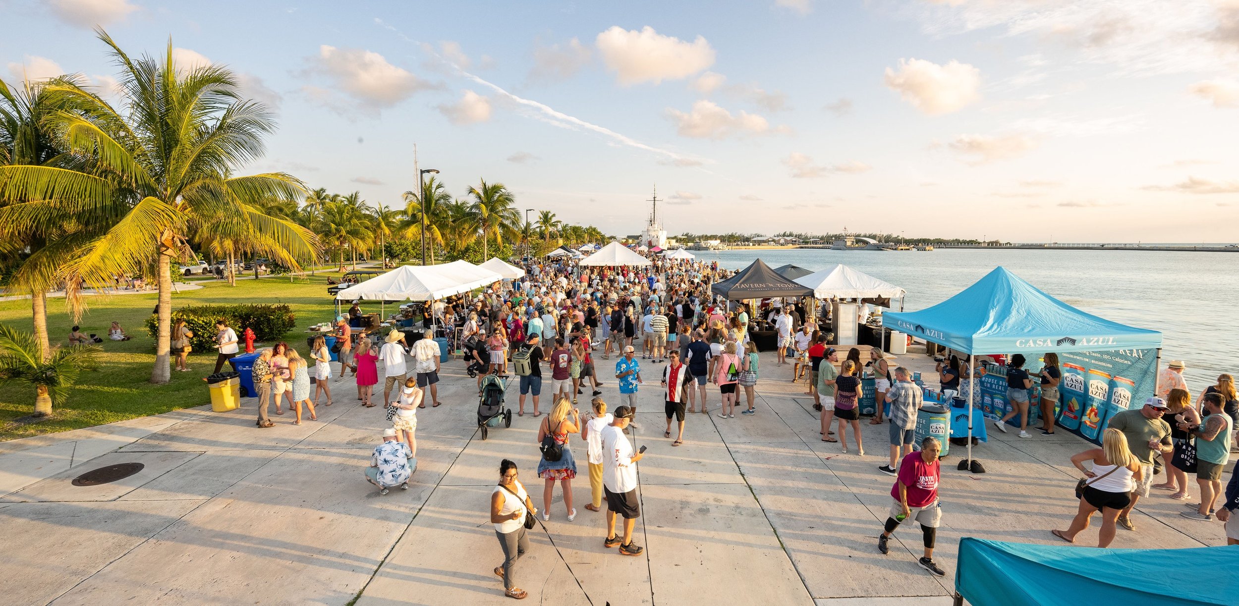 Crowd of people at an outdoor event near a waterfront with tents, palm trees, and a ship in the background during sunset.