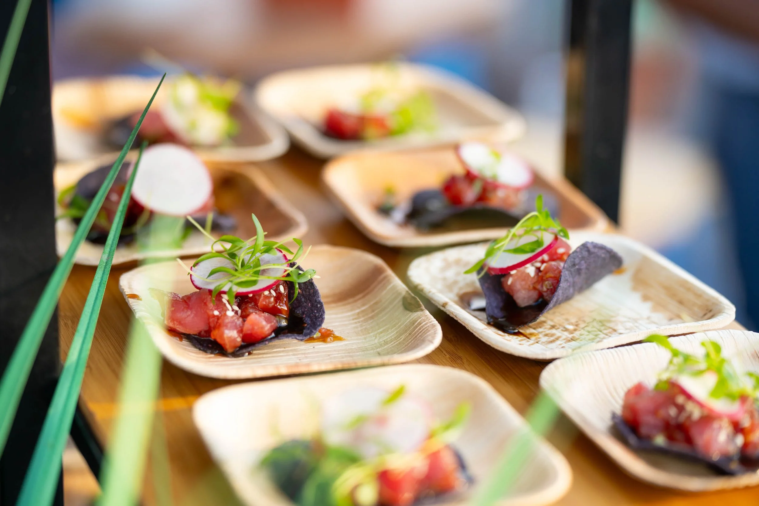 Close-up of small plates with gourmet sushi-style appetizers topped with radish slices and microgreens, displayed on a wooden surface at a food event.