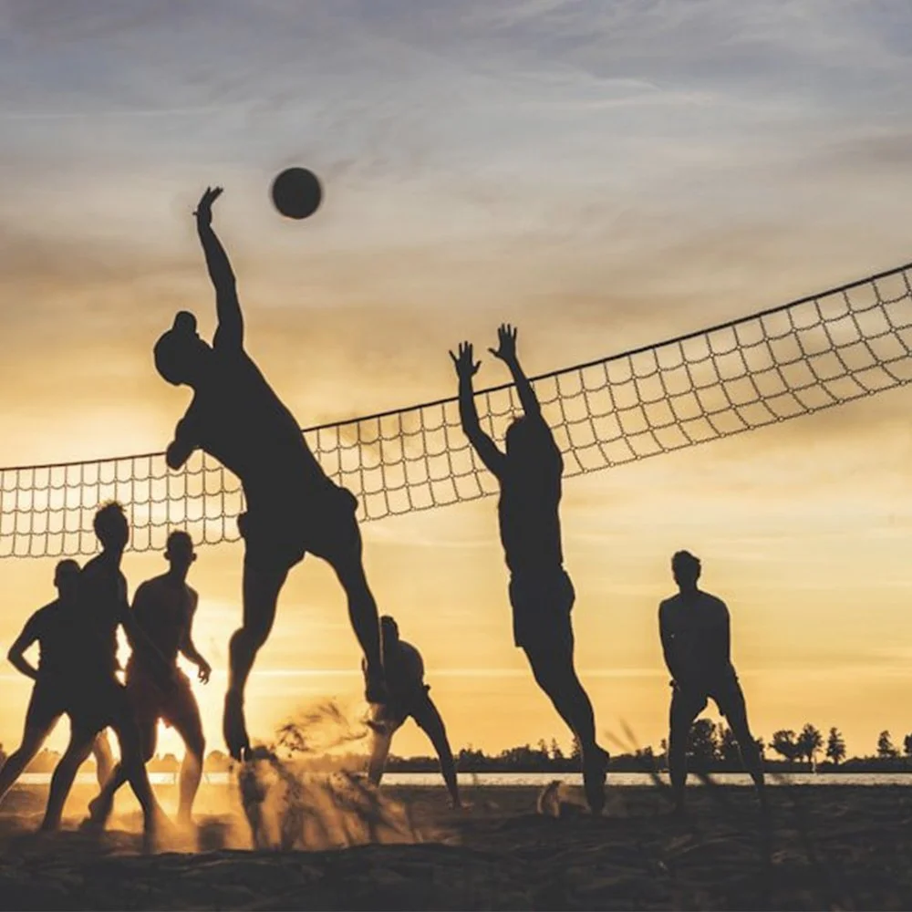 Silhouettes of six people playing beach volleyball at sunset, with two players jumping to hit the ball over the net.