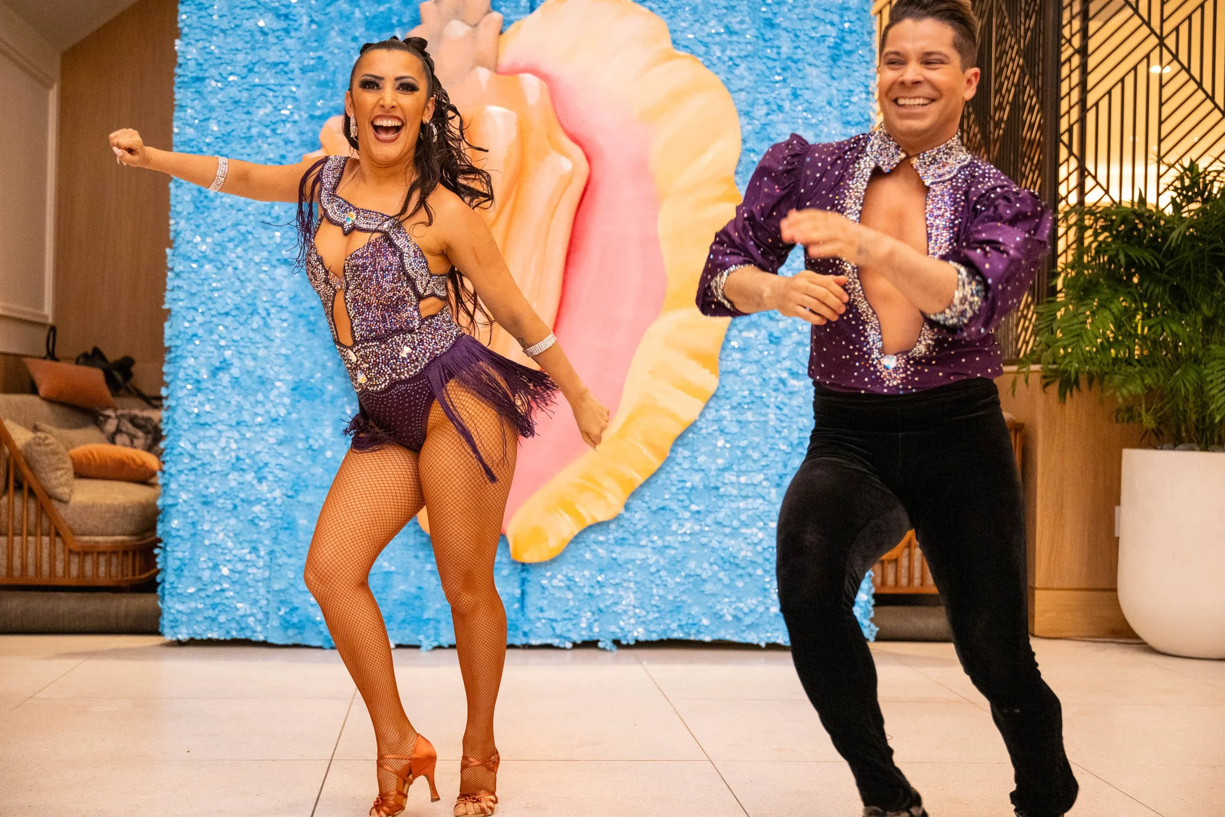 Two performers dancing in colorful costumes, with a blue textured backdrop and a large, pink, yellow, and orange flower decoration behind them, smiling and enjoying a lively performance.