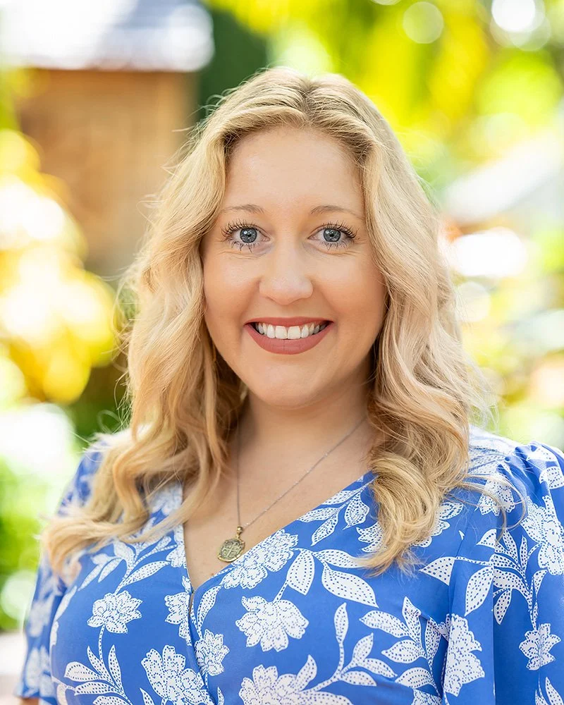 A woman with long blonde hair smiling outdoors with a blurred background of yellow and green foliage.