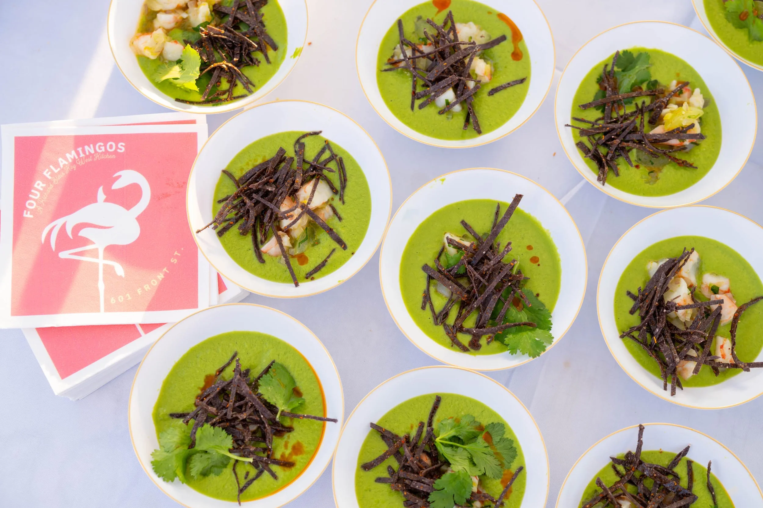 Several small bowls of green soup topped with shredded dark vegetables, possibly seaweed, and garnished with cilantro pieces. A pink card with a white flamingo logo and text reading 'Four Flamingos, 601 Front St.' is visible beside the bowls.