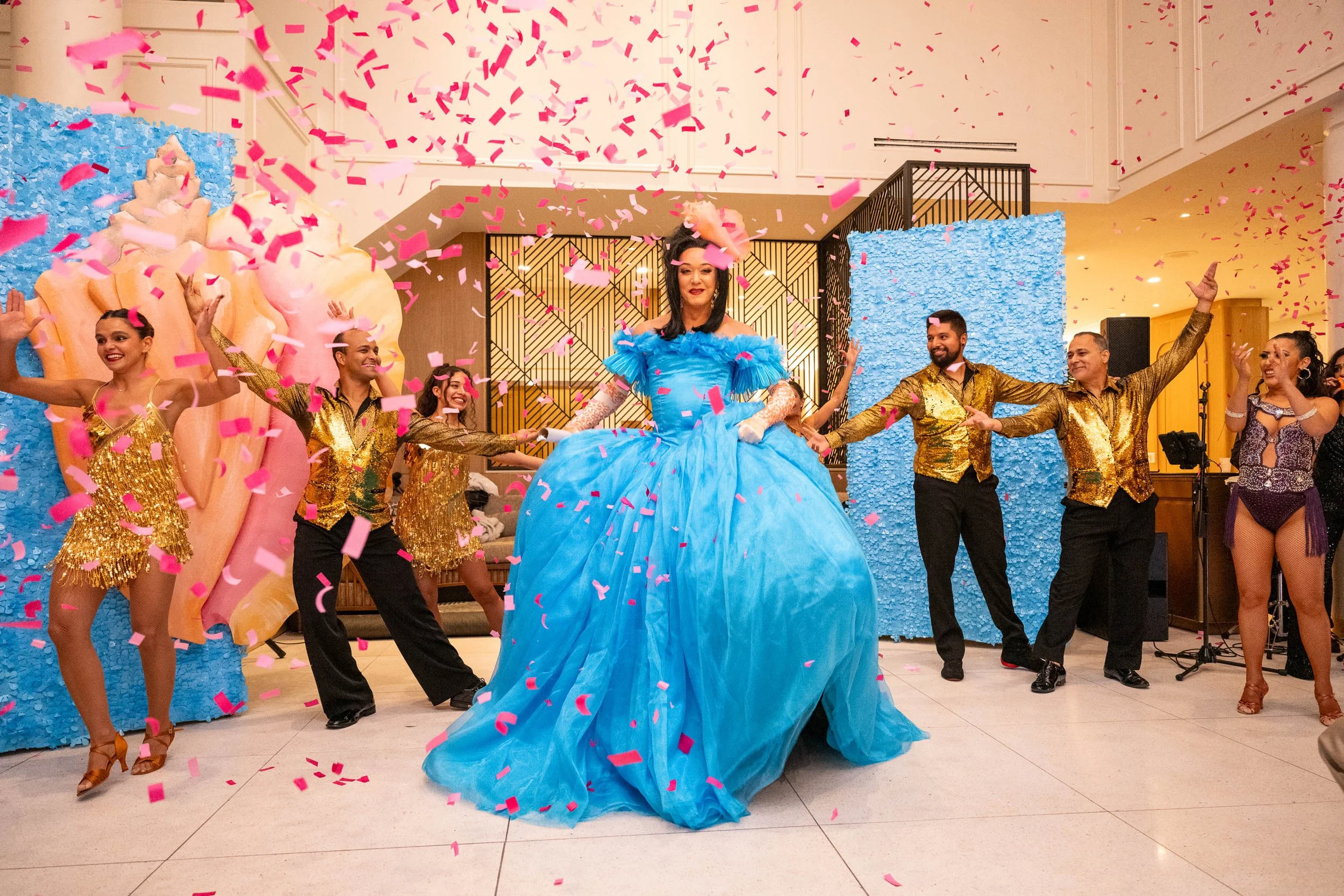 A group of performers celebrating with a woman dressed as a princess in a bright blue gown. Pink confetti falls around them, with some dancers in gold costumes and a woman in a showgirl outfit. The setting appears to be an indoor event or party with 