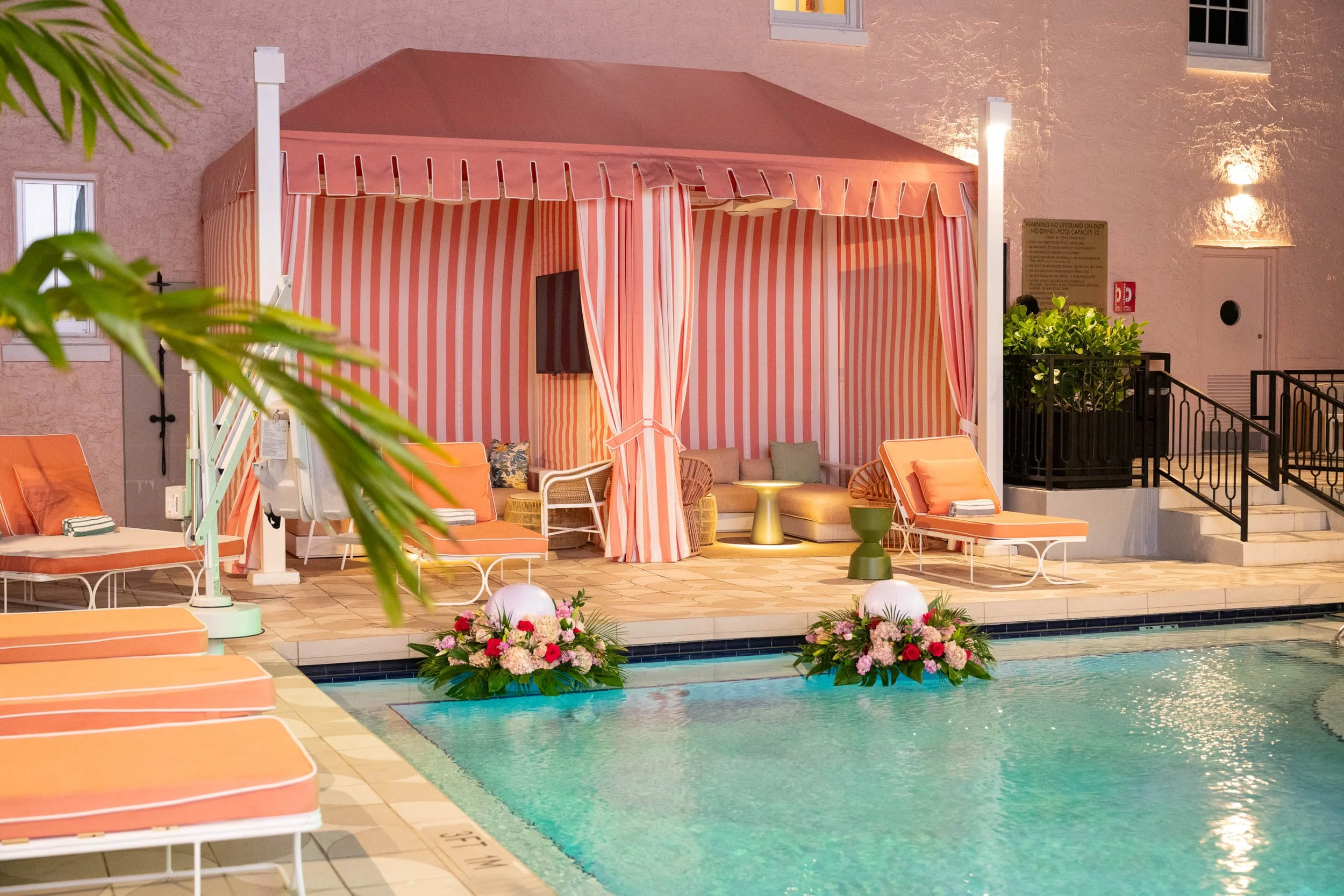Indoor pool area with pink and orange striped cabana, lounge chairs, tropical plants, and floral arrangements.