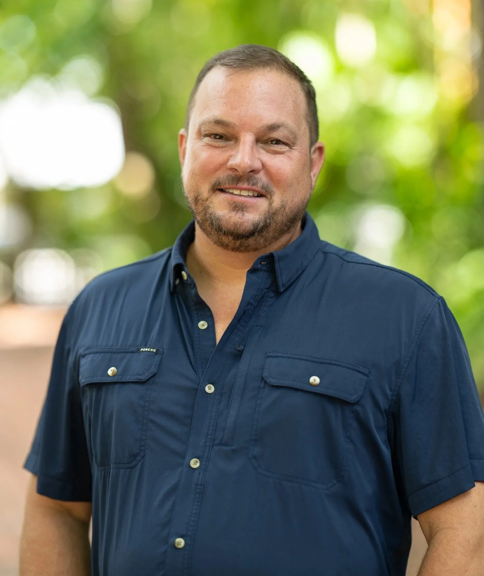 A man with short hair and a beard wearing a navy blue button-up shirt, standing outdoors with a blurred background of green trees.