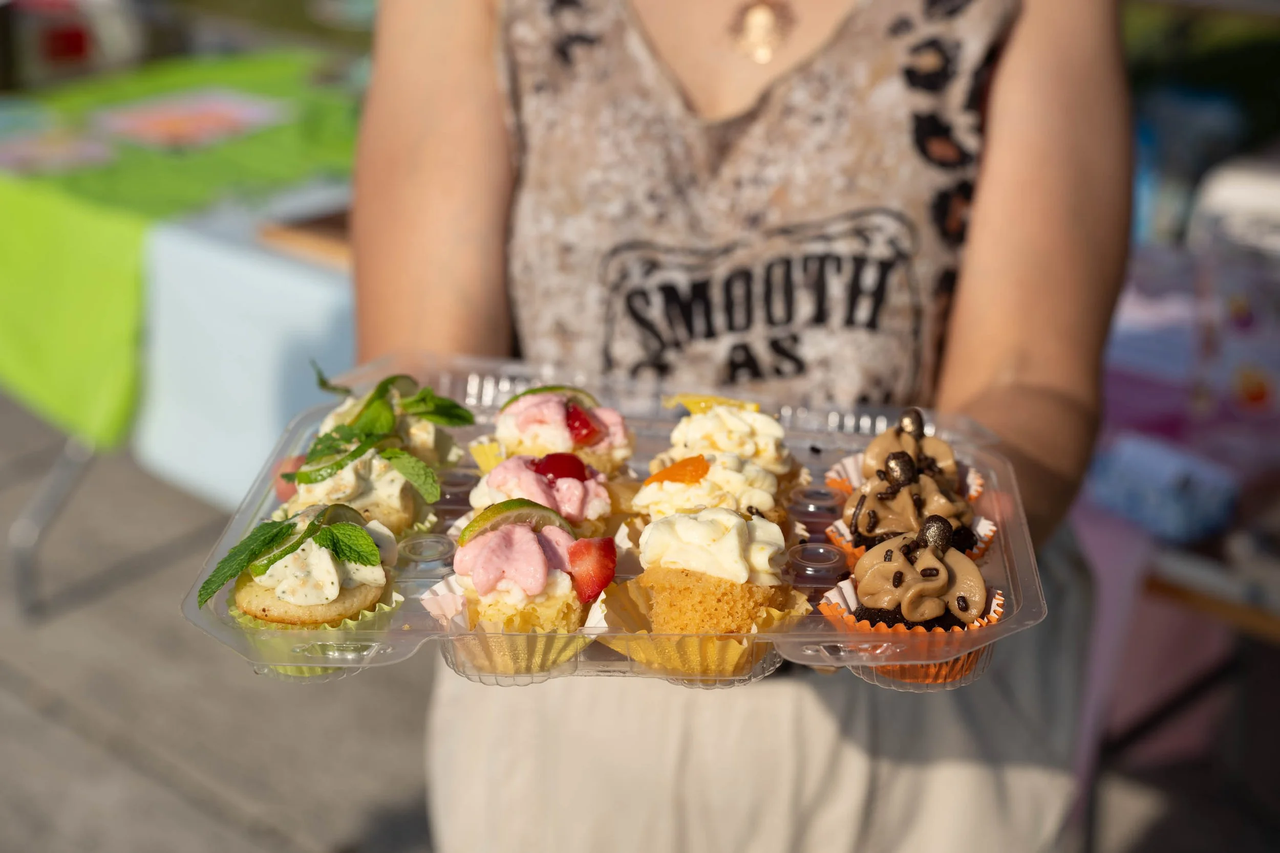 Person holding a clear plastic tray of assorted cupcakes at an outdoor market or event.