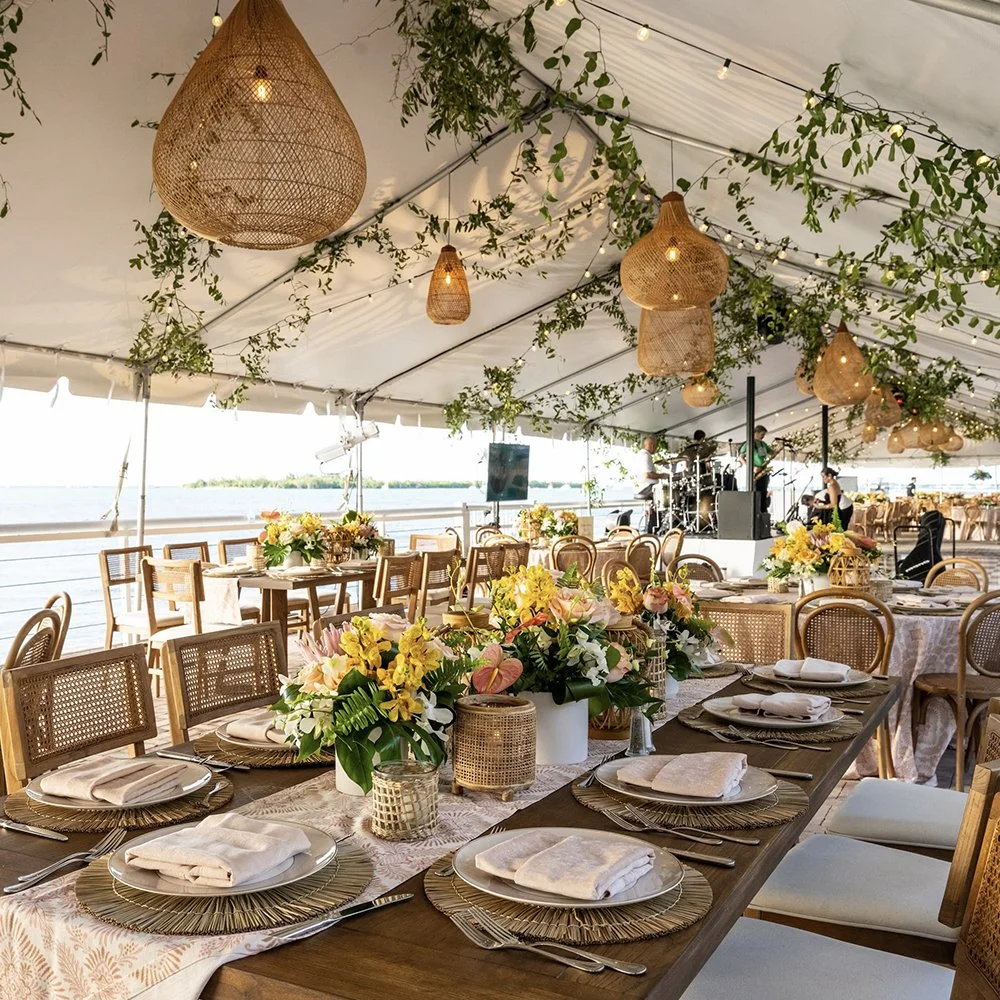 Elegant outdoor wedding reception under a tent by a body of water, decorated with hanging woven lanterns, greenery, and string lights, with a band playing live music in the background.