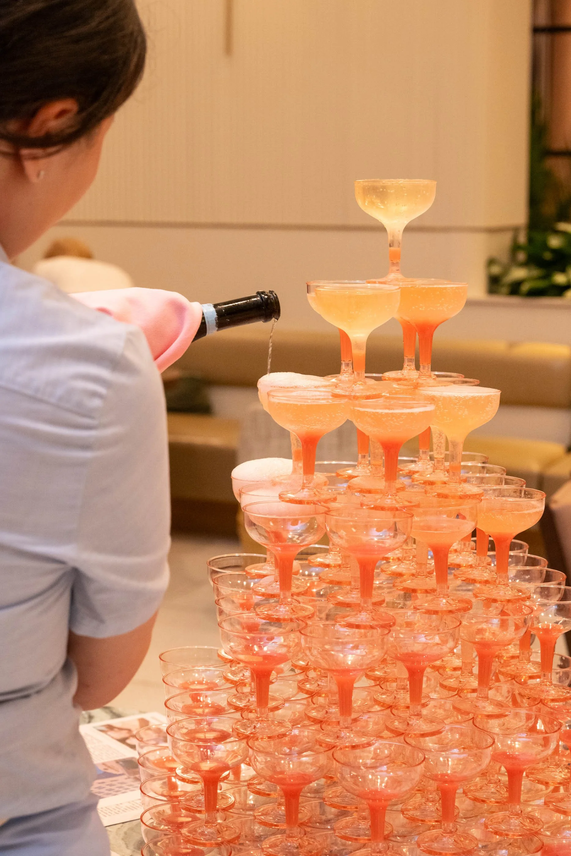 A person is pouring champagne into a pyramid of pink and orange cocktail glasses.