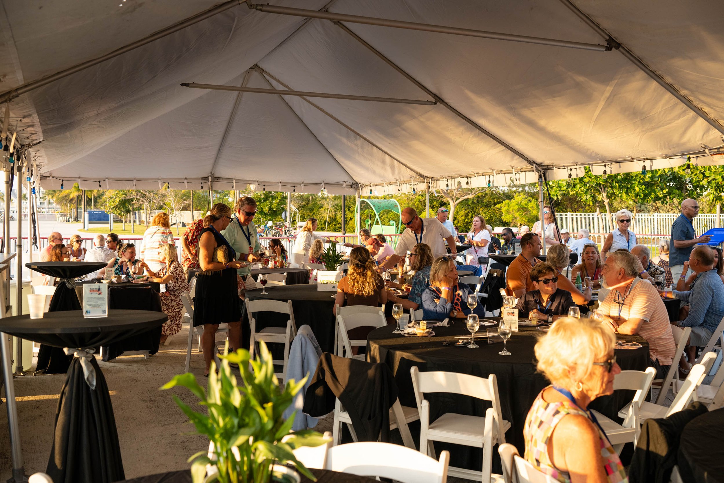 People dining under a large event tent, seated at tables with black tablecloths, engaged in conversations and enjoying drinks, with some standing and serving themselves, outdoors on a sunny day.