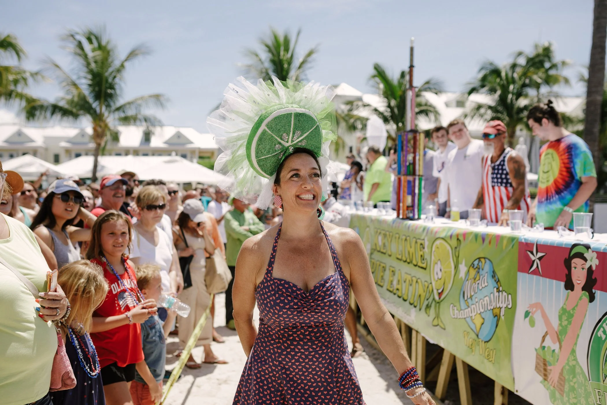 A woman wearing a lime slice hat, smiling at a tropical outdoor event with palm trees and a crowd of people in the background.