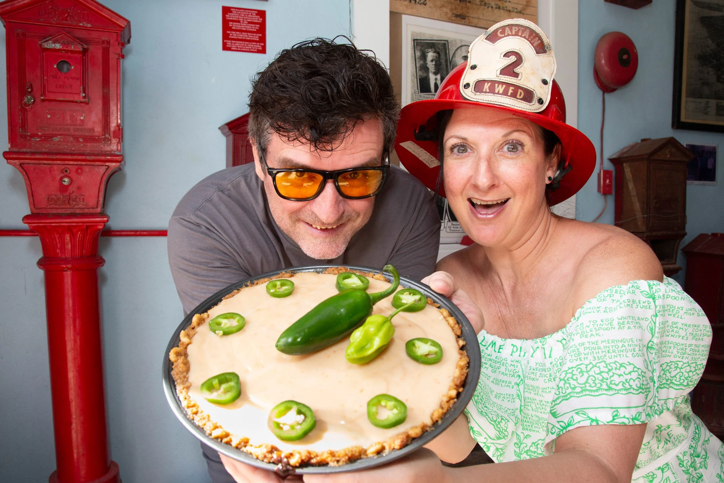 A man and woman dressed as a firefighter holding a pie decorated with jalapeño peppers, with the woman wearing a firefighter helmet.
