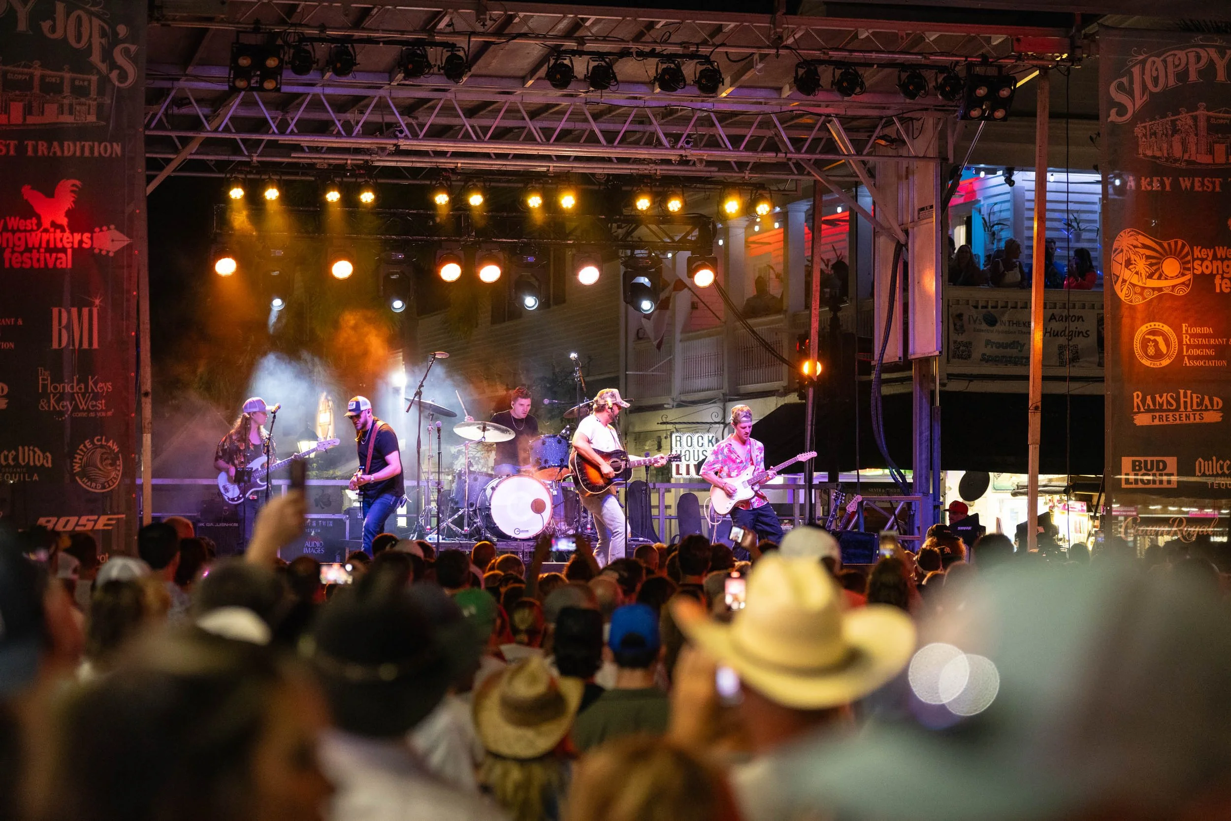 Live band performing on stage at night with audience in front, stage with lighting, and banners on both sides advertising various sponsors.