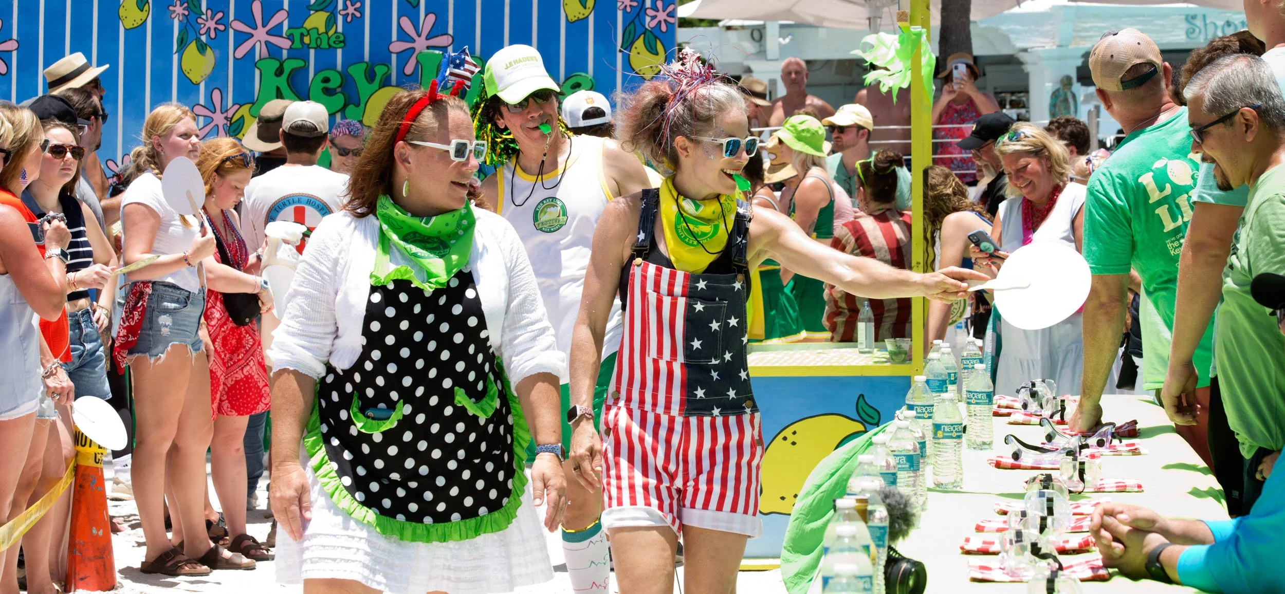People participating in a summer contest or event outdoors, some wearing playful patriotic-themed clothing, with a colorful, beach-themed backdrop and a table with bottled water and prizes.