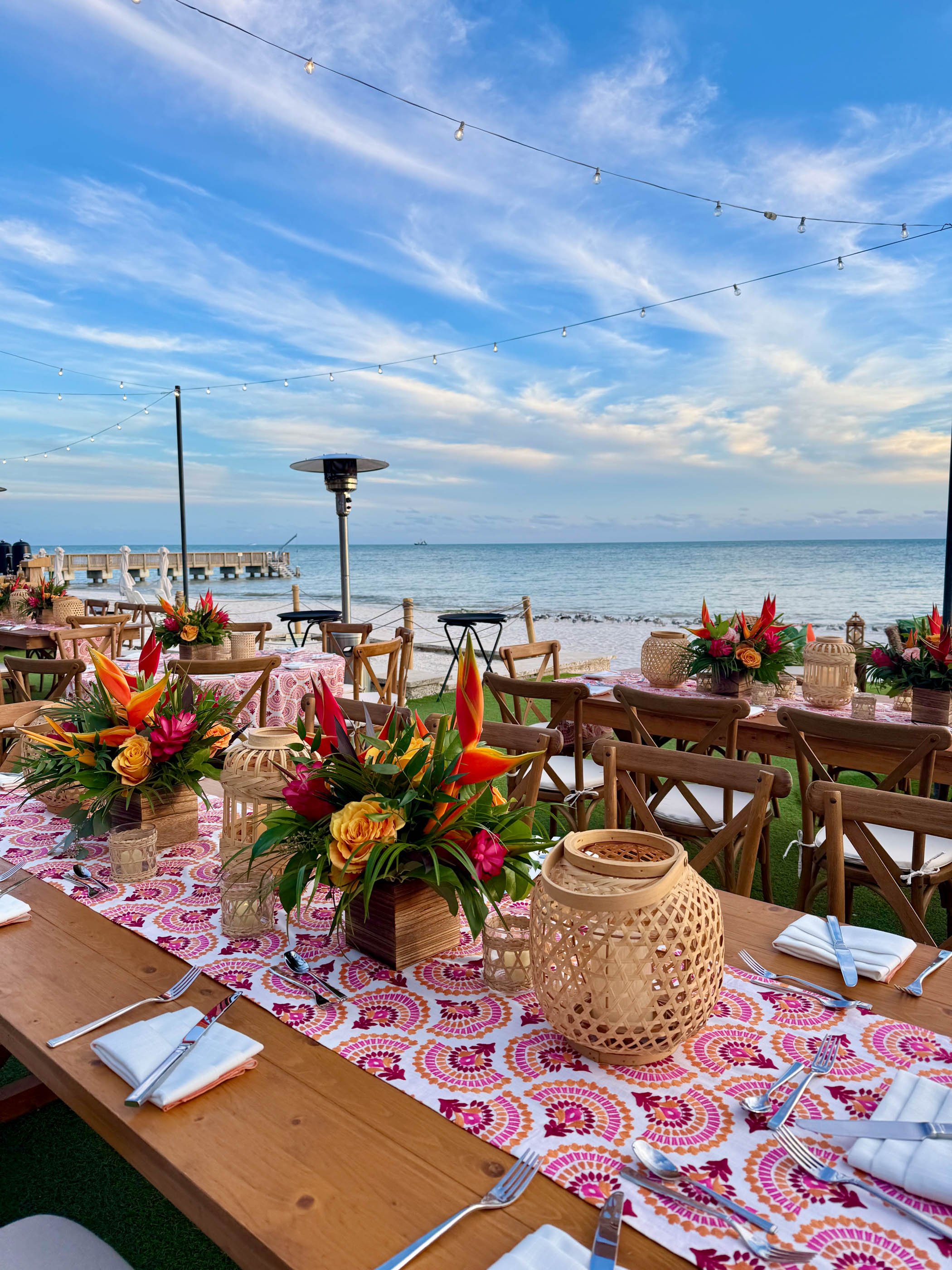 Outdoor seaside dining setup with a long wooden table decorated with floral centerpieces, lanterns, and arranged with silverware and white napkins, overlooking the ocean under string lights and a partly cloudy sky.