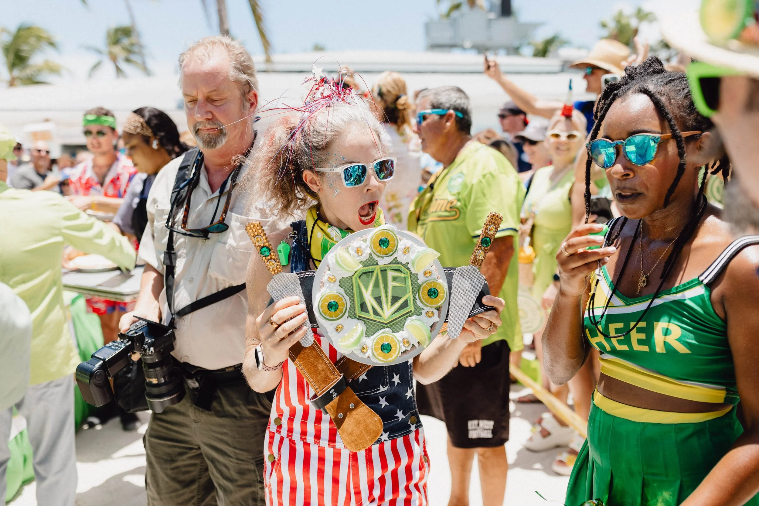 Crowd of people at a celebration, woman in the center wearing sunglasses and colorful clothing holding a decorative cake, woman on the right in a green cheerleader outfit, bright outdoor setting.