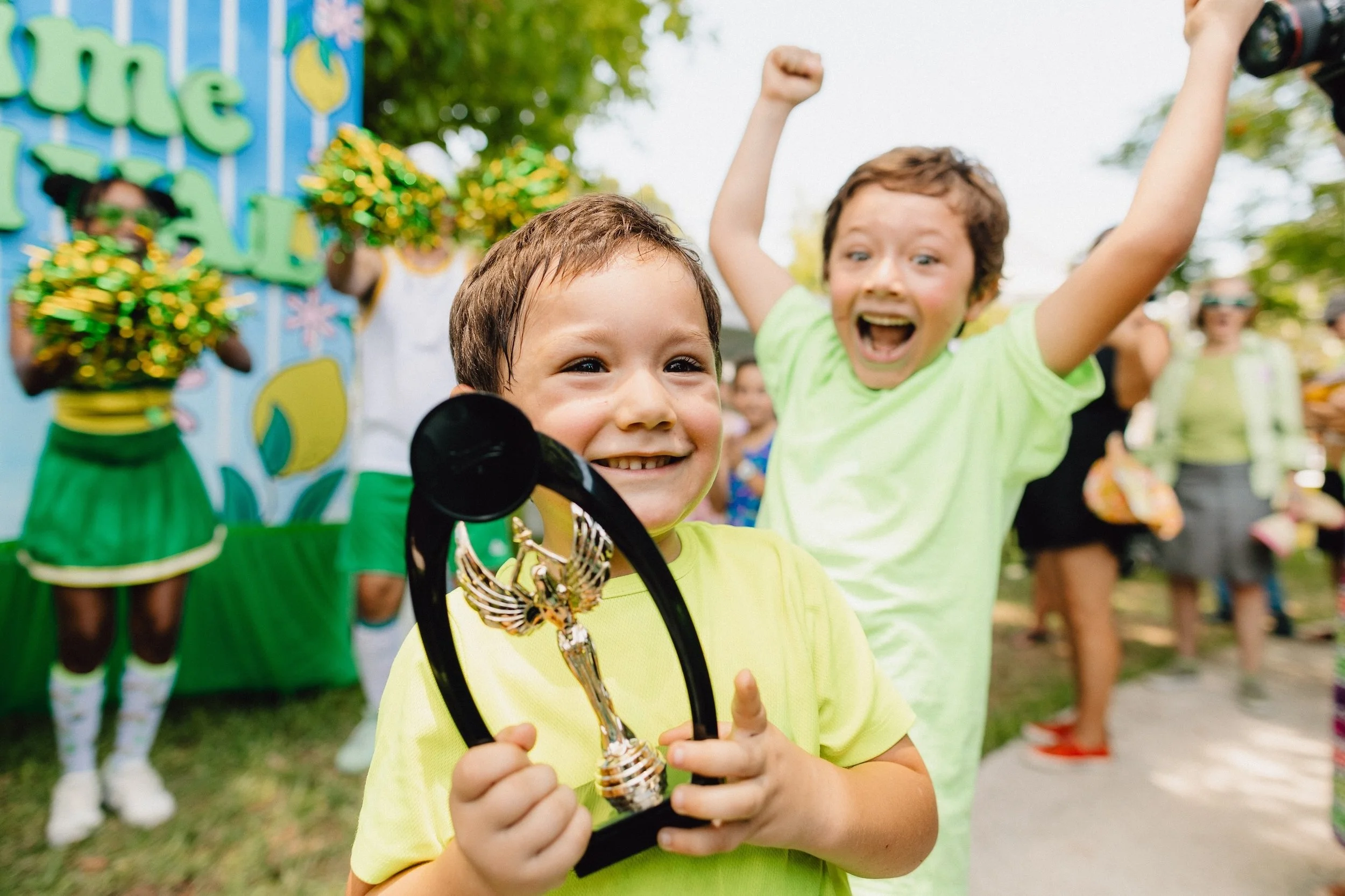 A young boy smiling and holding a trophy at an outdoor event, with another boy celebrating in the background, surrounded by other children and cheerleaders.