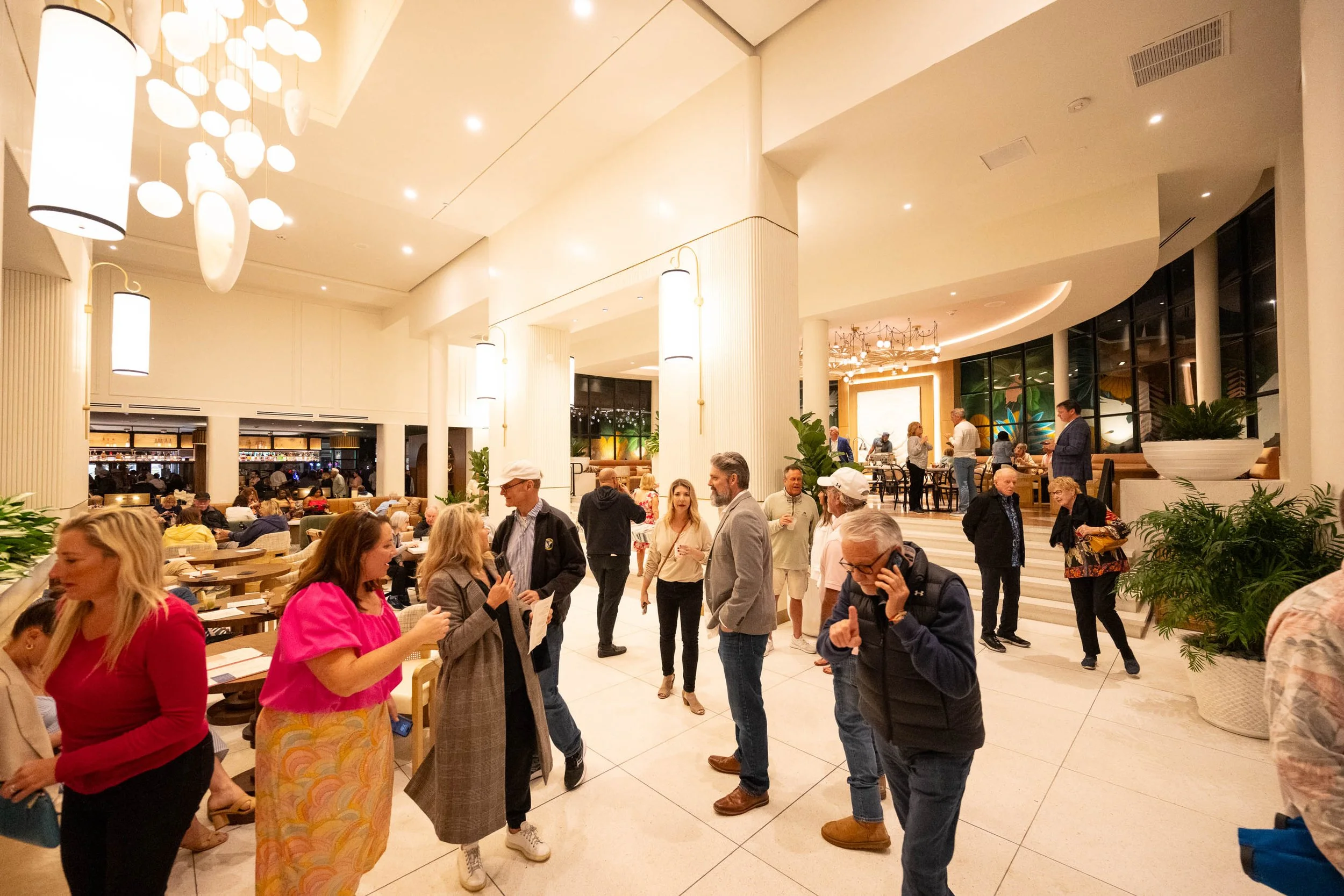 A lively indoor scene at a social gathering or restaurant with people talking and mingling. The space has bright lighting, modern decor, and large windows. Potted plants and elegant furniture enhance the atmosphere.