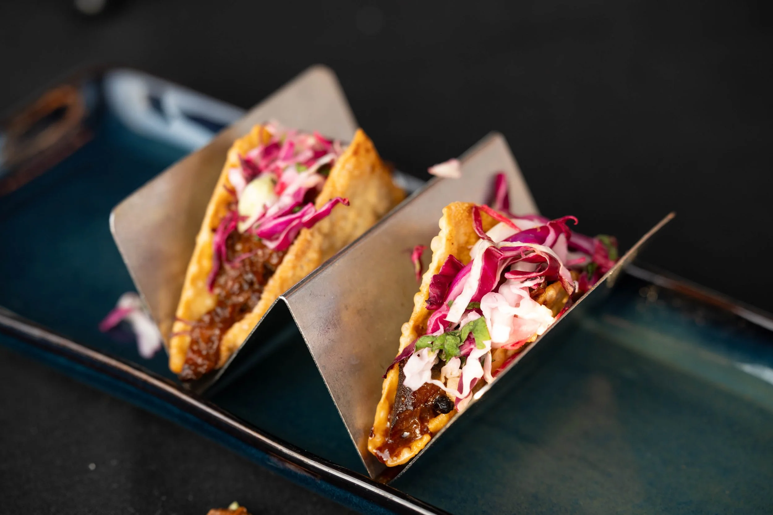 Two tacos filled with beef and topped with shredded red and white cabbage, served on a metal taco holder