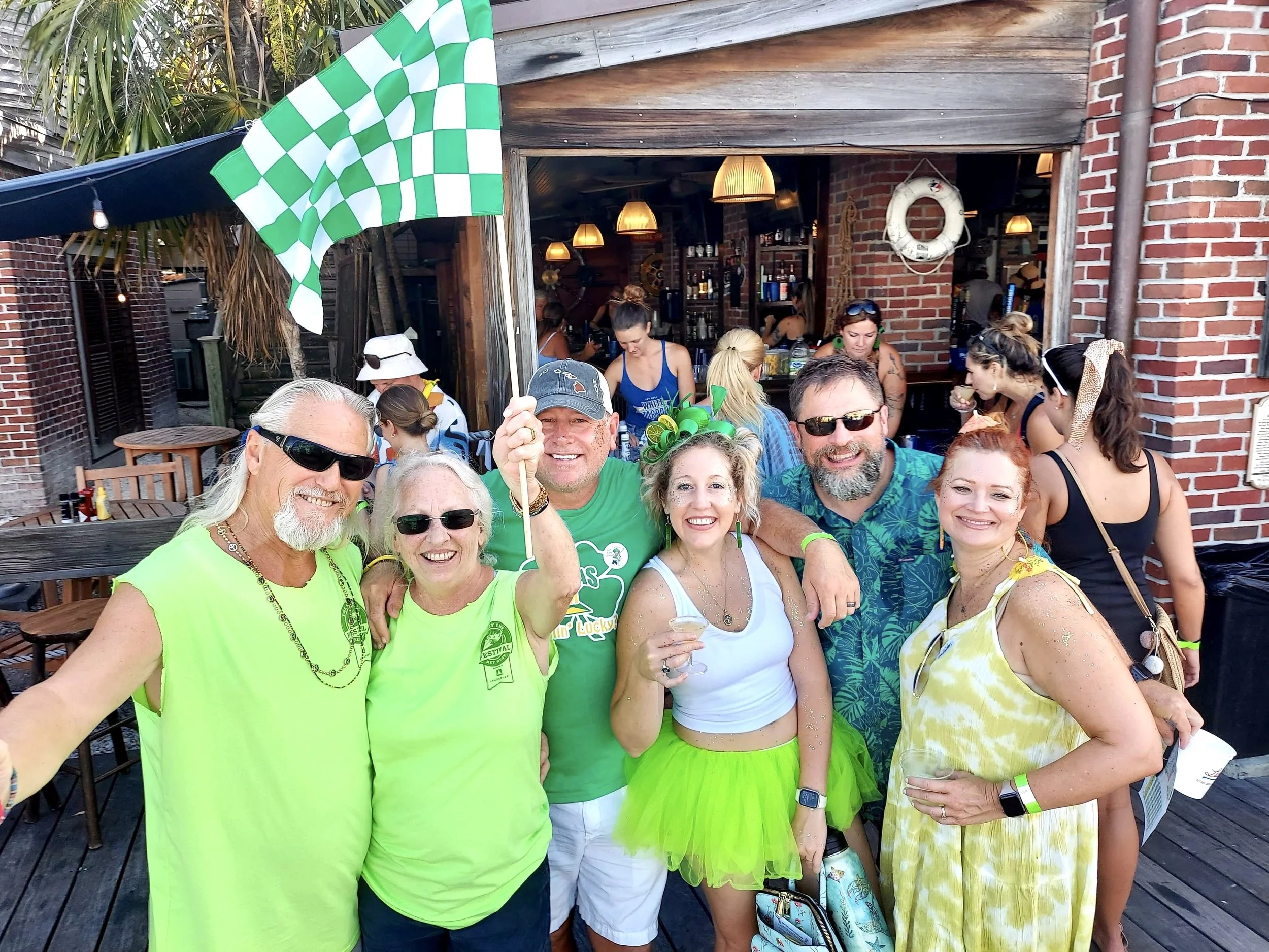 Group of seven people celebrating outdoors in front of a brick bar, with some wearing bright green and yellow clothing, holding drinks, smiling, and enjoying a sunny day.