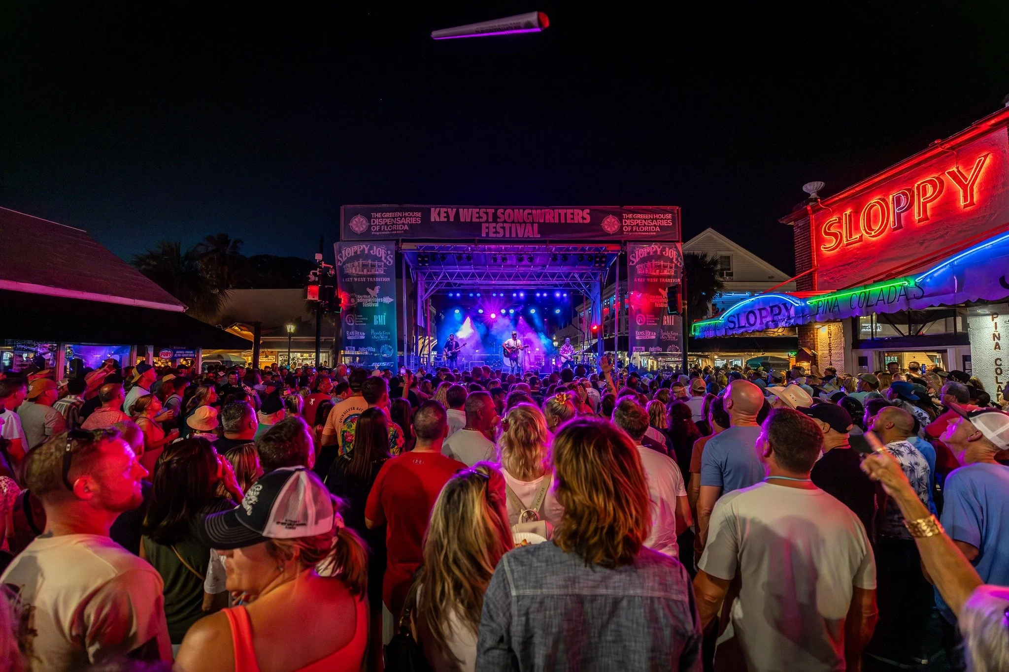 A crowd of people at night watching a live band perform on an outdoor stage during a festival, with colorful lighting and neon signs.