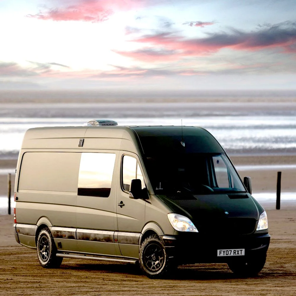 A black and beige camper van parked on a sandy beach with the ocean and a colorful sunset sky in the background.