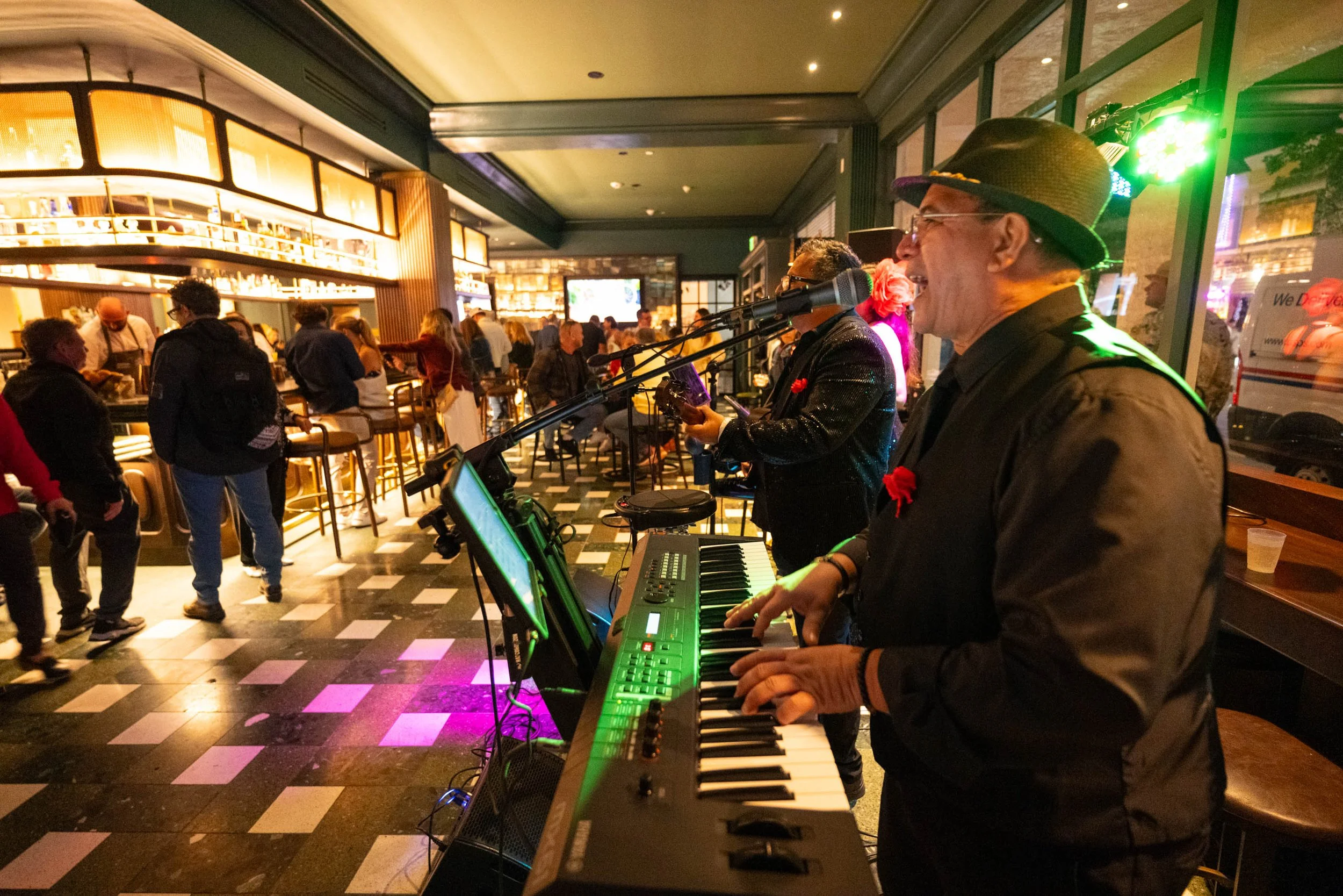 Musicians performing at a lively indoor restaurant bar, with the man playing piano and singing into a microphone, surrounded by customers and staff, bar shelves in the background, warm lighting, and a checkered floor.