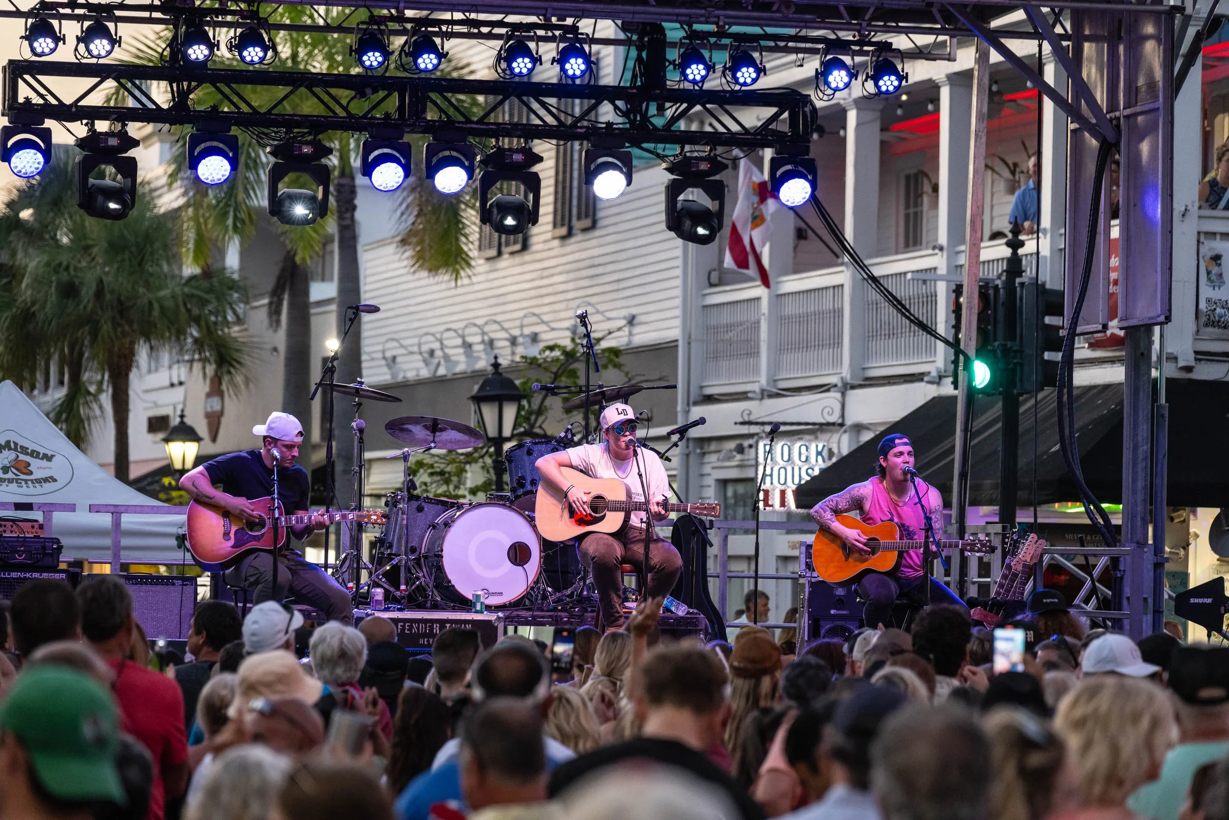 A live outdoor music performance on a stage with three musicians playing guitars and singing, surrounded by a large crowd.